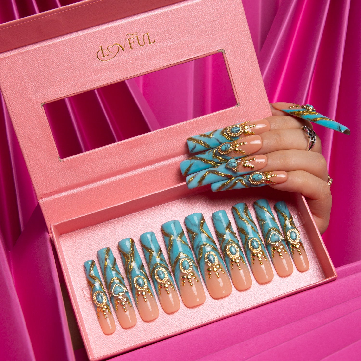 Hand wearing turquoise and gold press-on acrylic nails, displayed above matching nail set in pink Lovful branded box. Nails feature intricate golden decor and sparkling gems, set against a pink fabric backdrop.