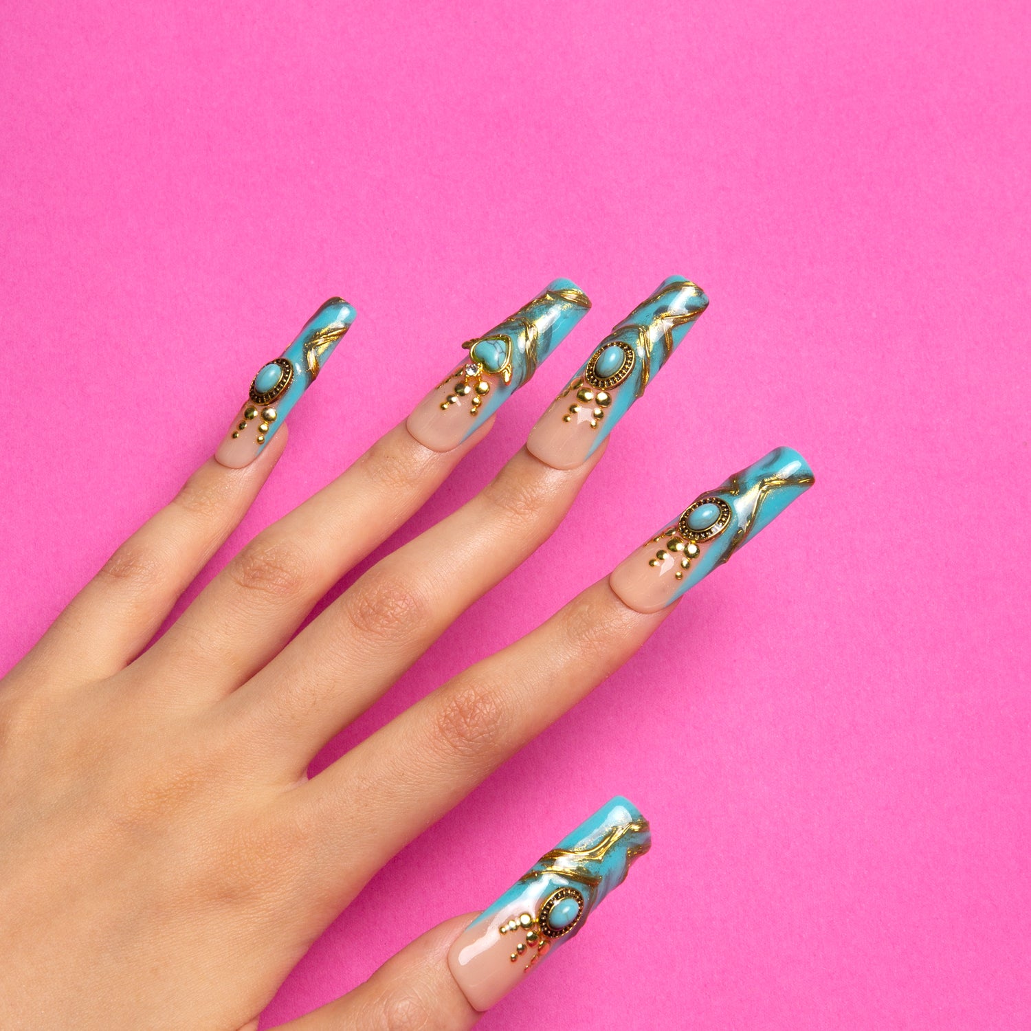 Hand with long square-shaped press-on nails adorned with luxurious turquoise and gold decor, featuring intricate golden patterns and sparkling gems, on a pink background. Perfect for glamorous occasions.