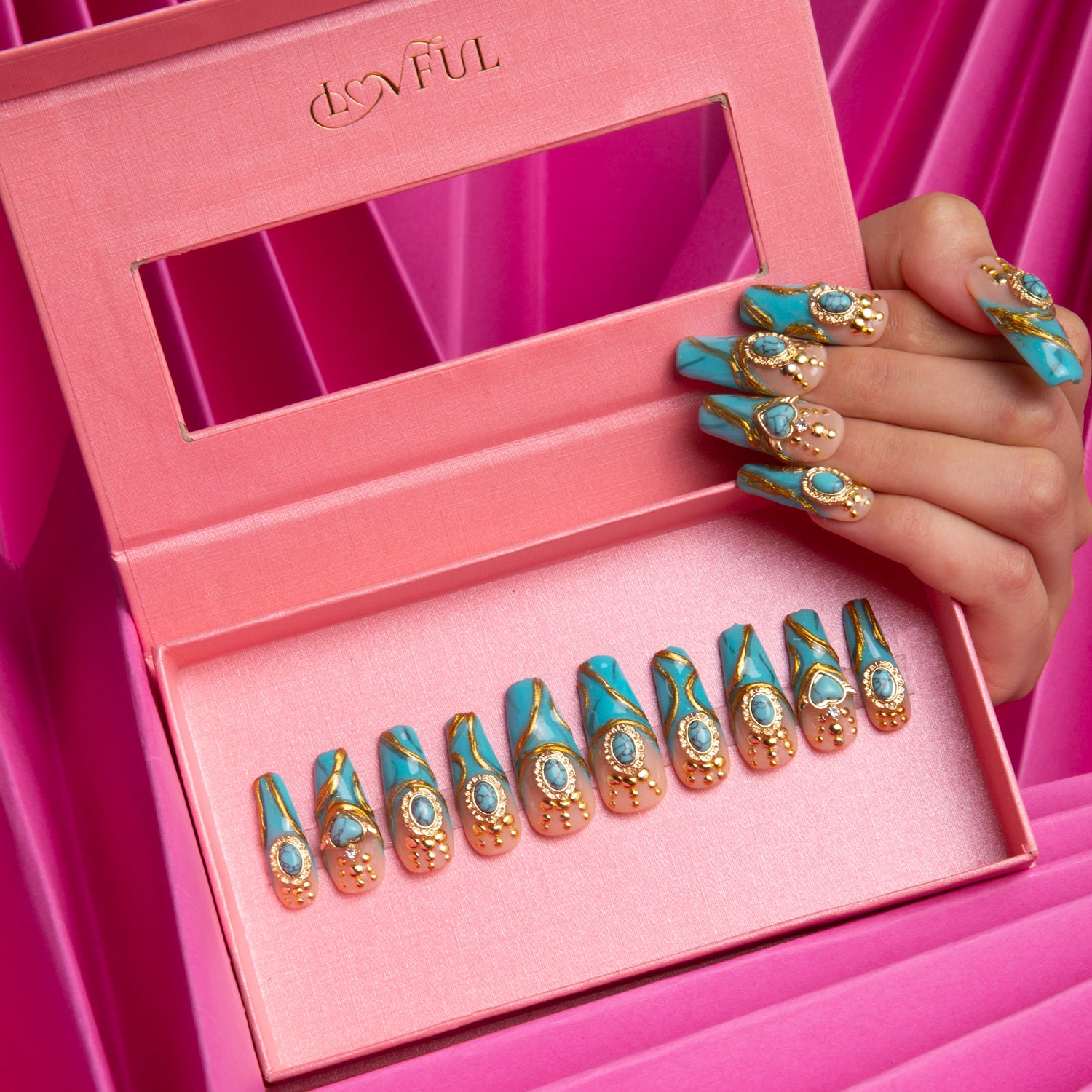 Turquoise coffin press-on nails with golden decor and sparkling gems in a pink box, held by a hand with matching nails, against a vibrant pink background. Lovful branded box.