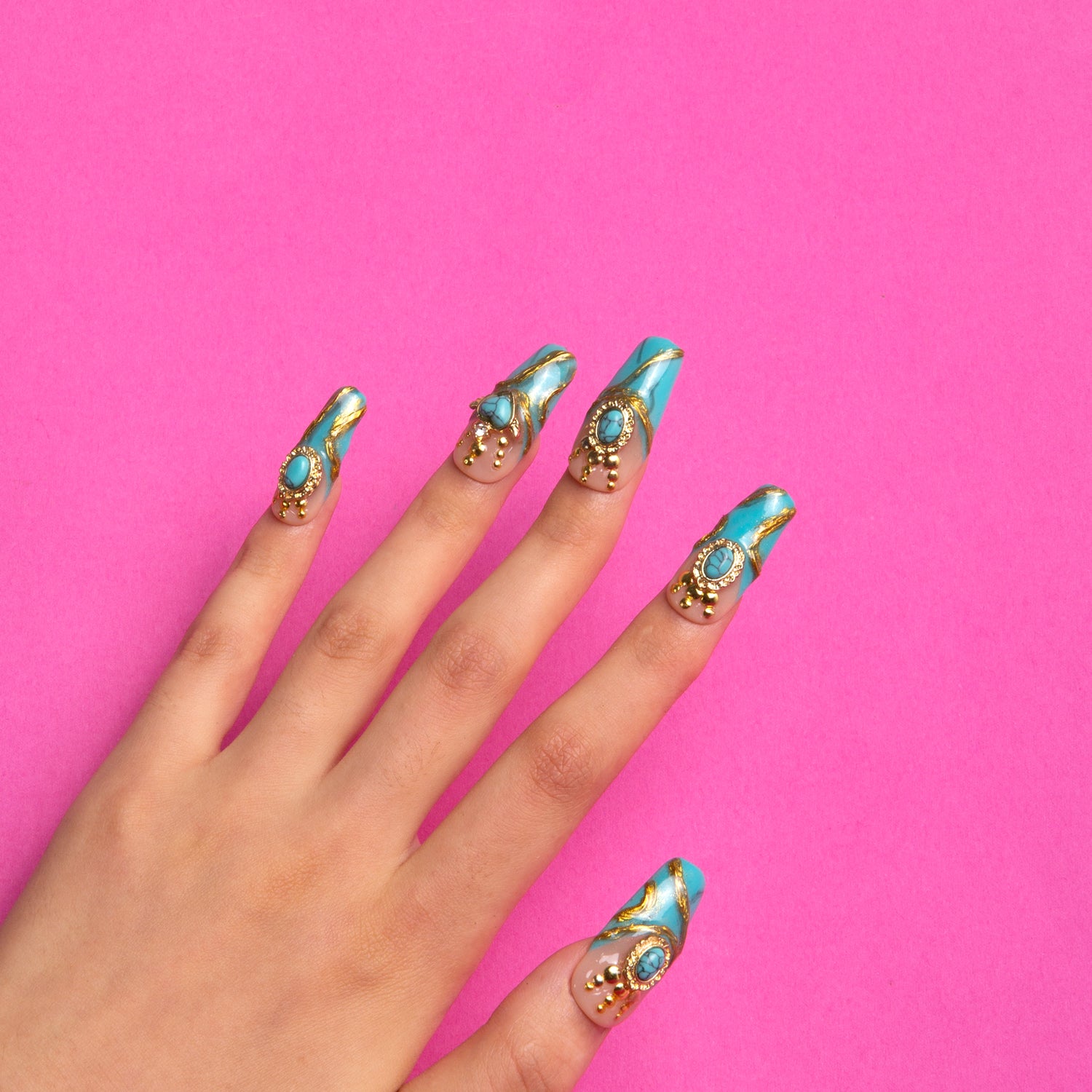 Hand model displaying luxurious turquoise coffin-shaped press-on nails adorned with golden decor and sparkling gems against a pink background.