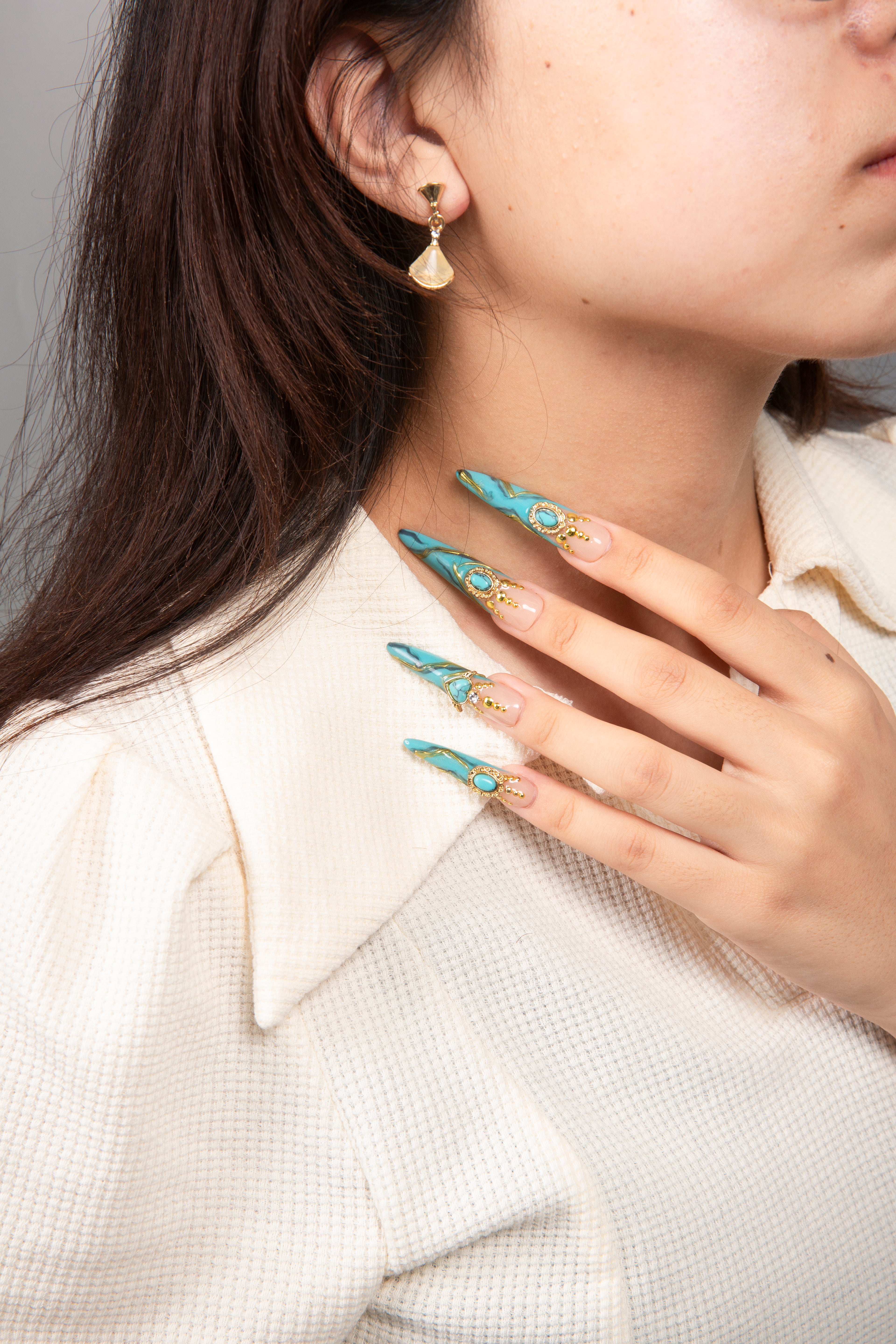 Close-up of woman's hand with Lovful's H122 Turquoise press-on nails, featuring turquoise nails with golden decor and sparkling gems, against a white textured blouse.