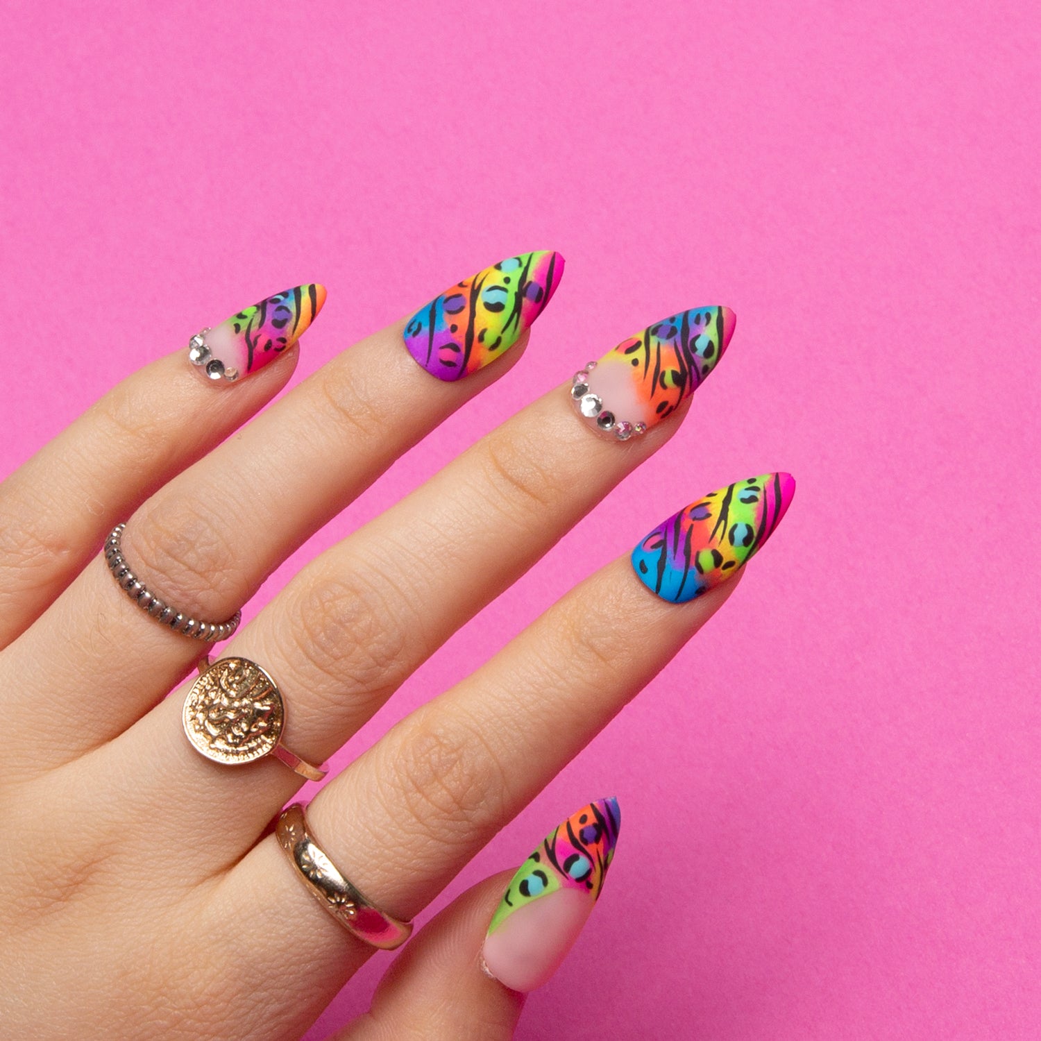 Hand displaying almond-shaped press-on nails with colorful French tips and leopard prints, accented with rings on a pink background. Perfect for adding a playful and confident touch.