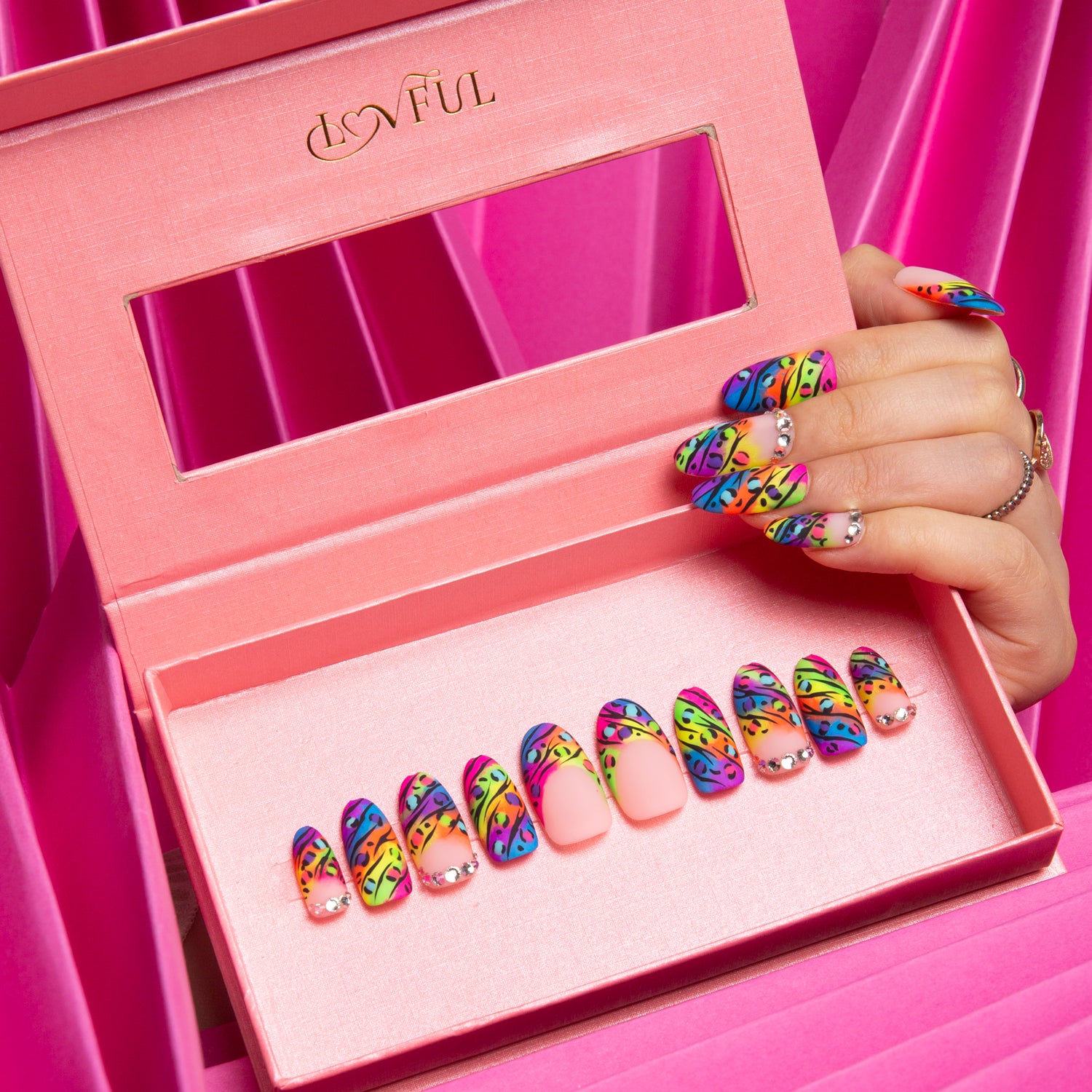 Pink box displaying a set of colorful press-on acrylic nails with bright French tips and leopard prints, matching hand holding the box against pink fabric drapes