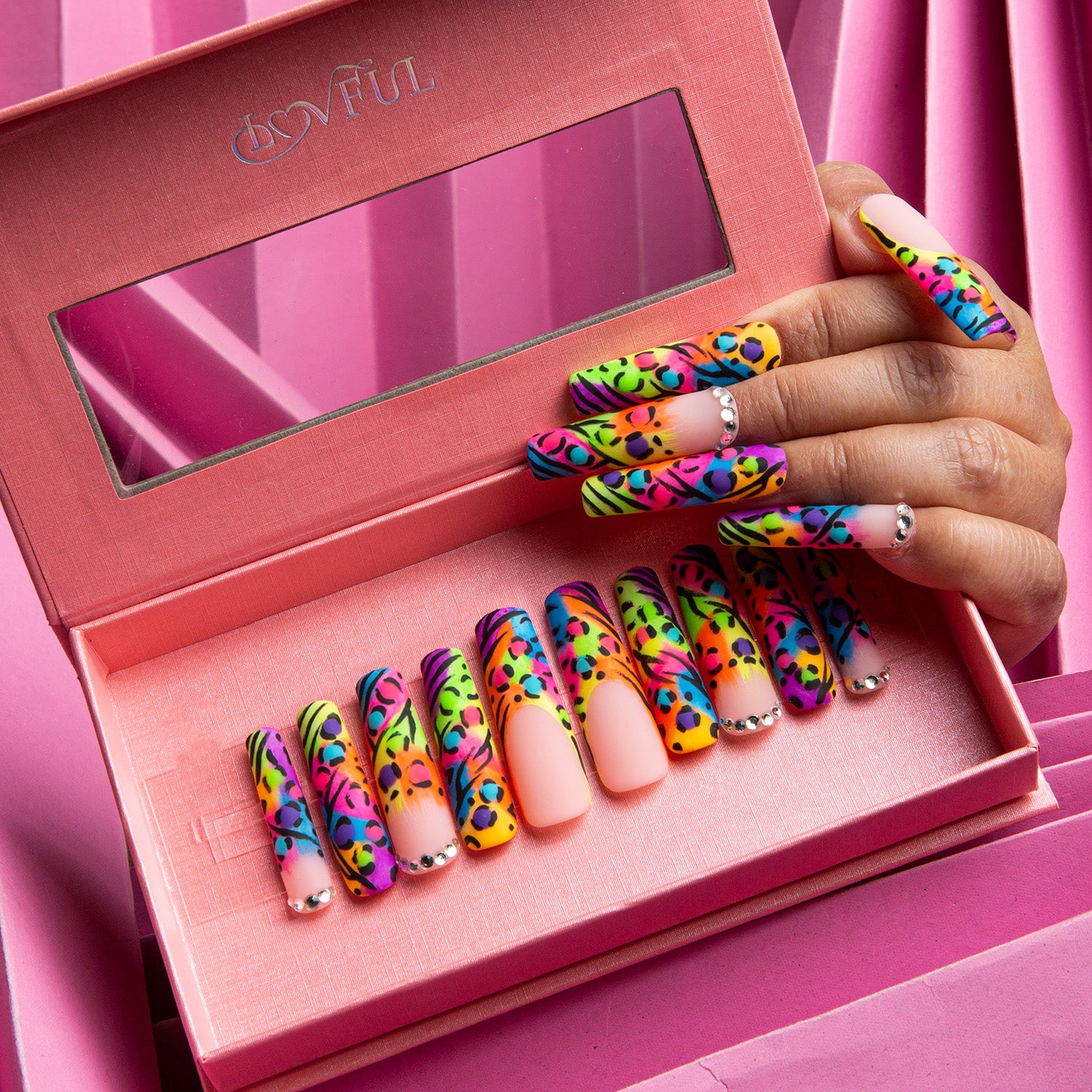 Colorful press-on nails with leopard print French tips displayed in a pink box, hand showing applied nails, vibrant pink background.