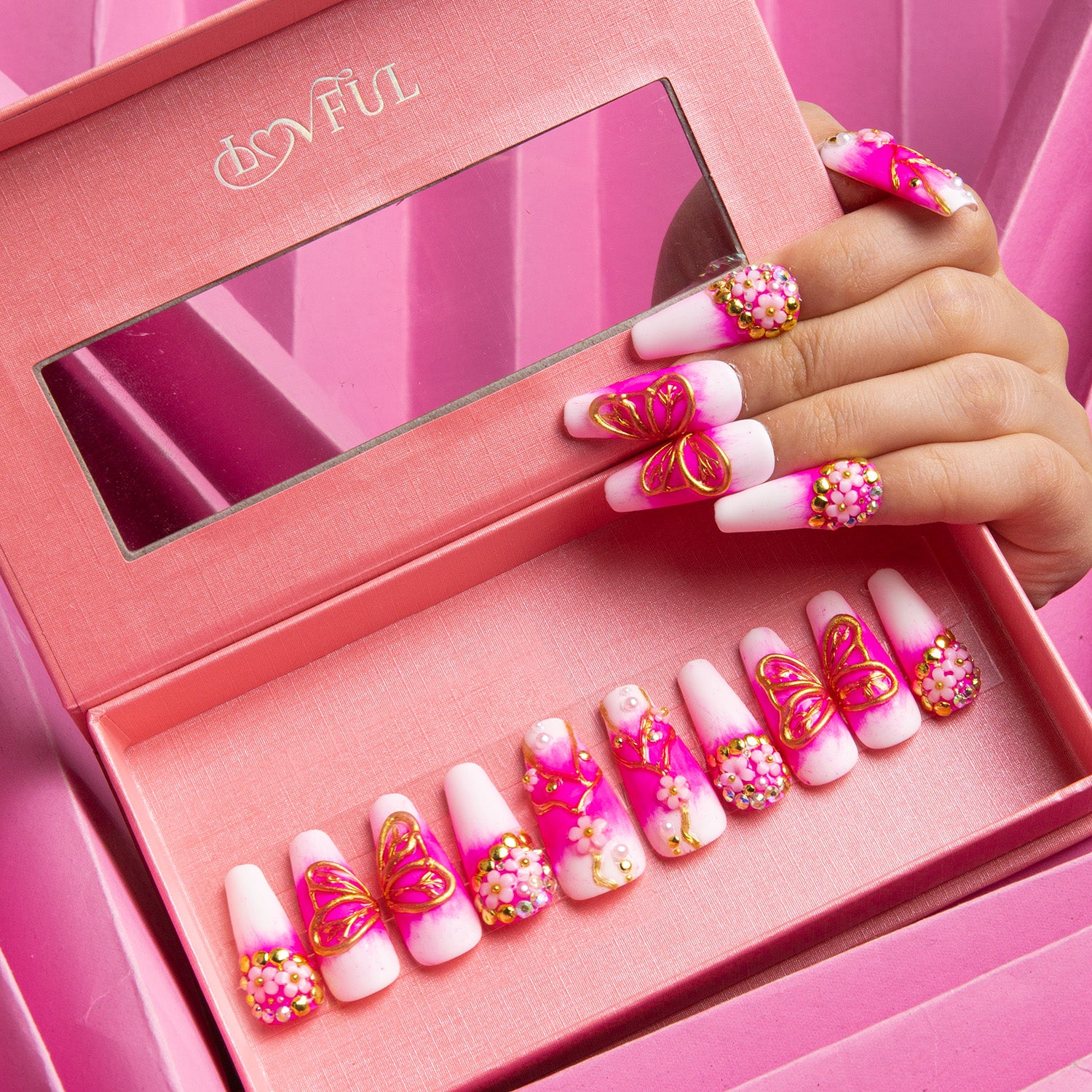 Set of press-on acrylic nails with white base, bright pink accents, and golden butterflies displayed in a pink Lovful box with built-in mirror. Hand with some nails applied also shown.