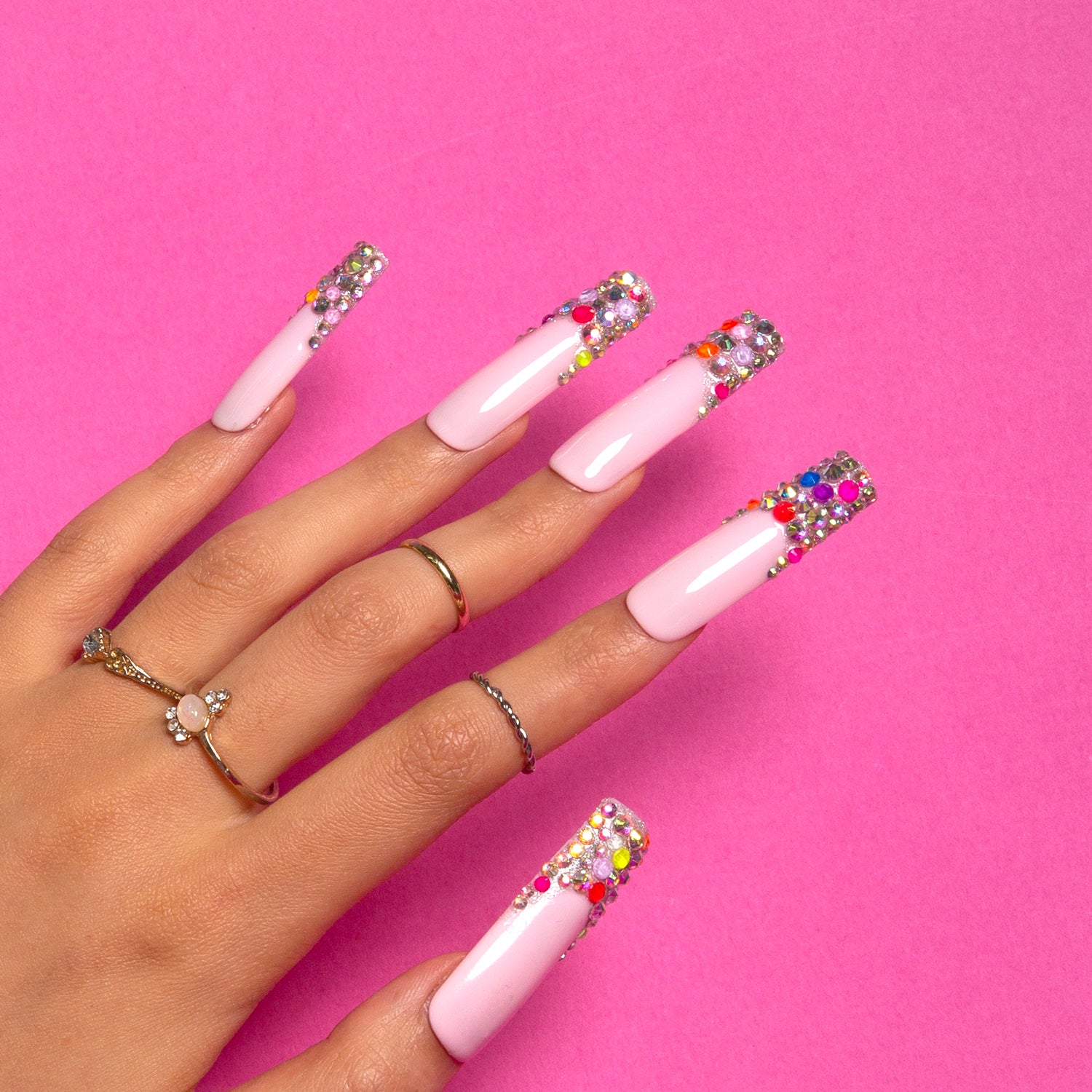 Hand with Jelly Bean Press-On Nails showcasing nude base with colorful, sugary bean accents on tips against pink background, wearing multiple rings