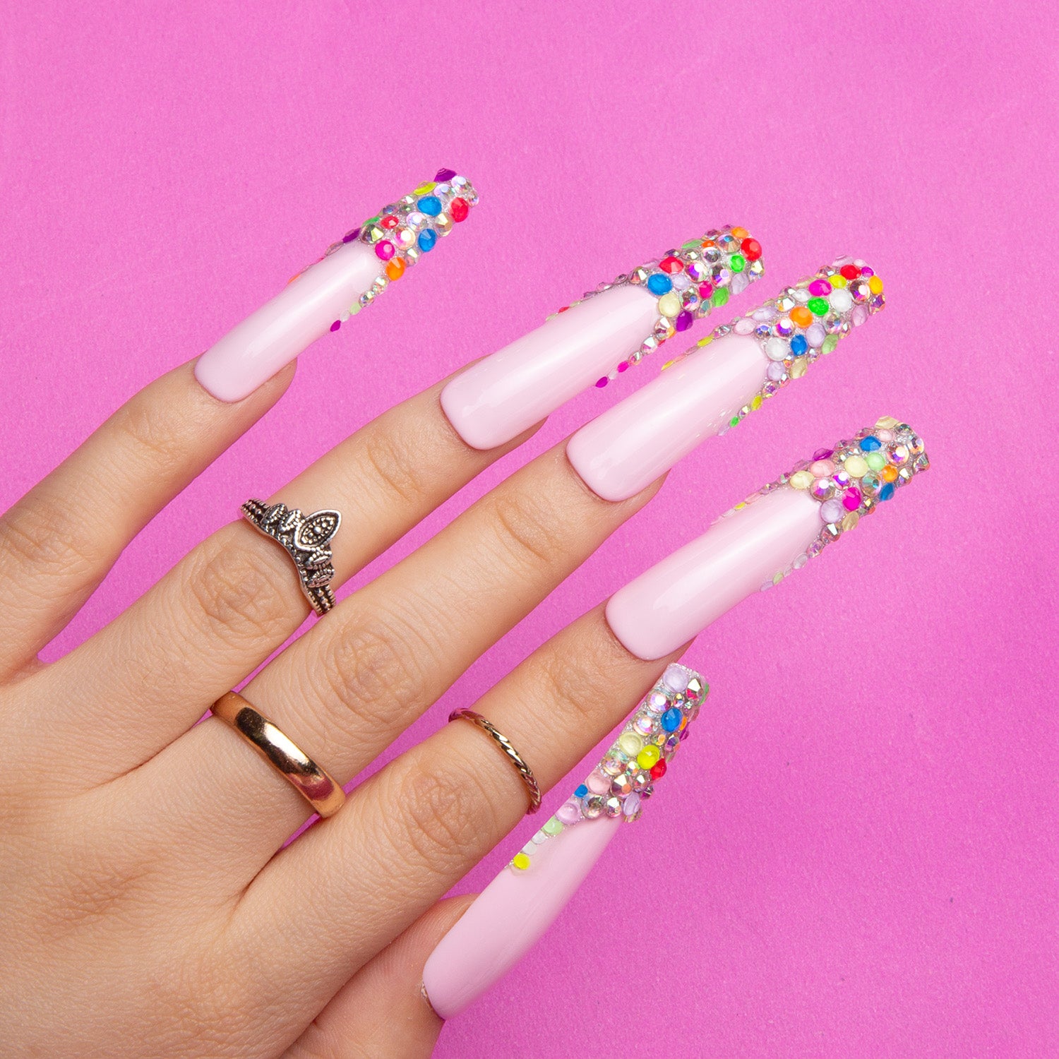 Coffin-shaped press-on nails with nude base and colorful jelly bean design tips, featuring small, colorful sugary beads. Hand wearing rings in front of pink background.