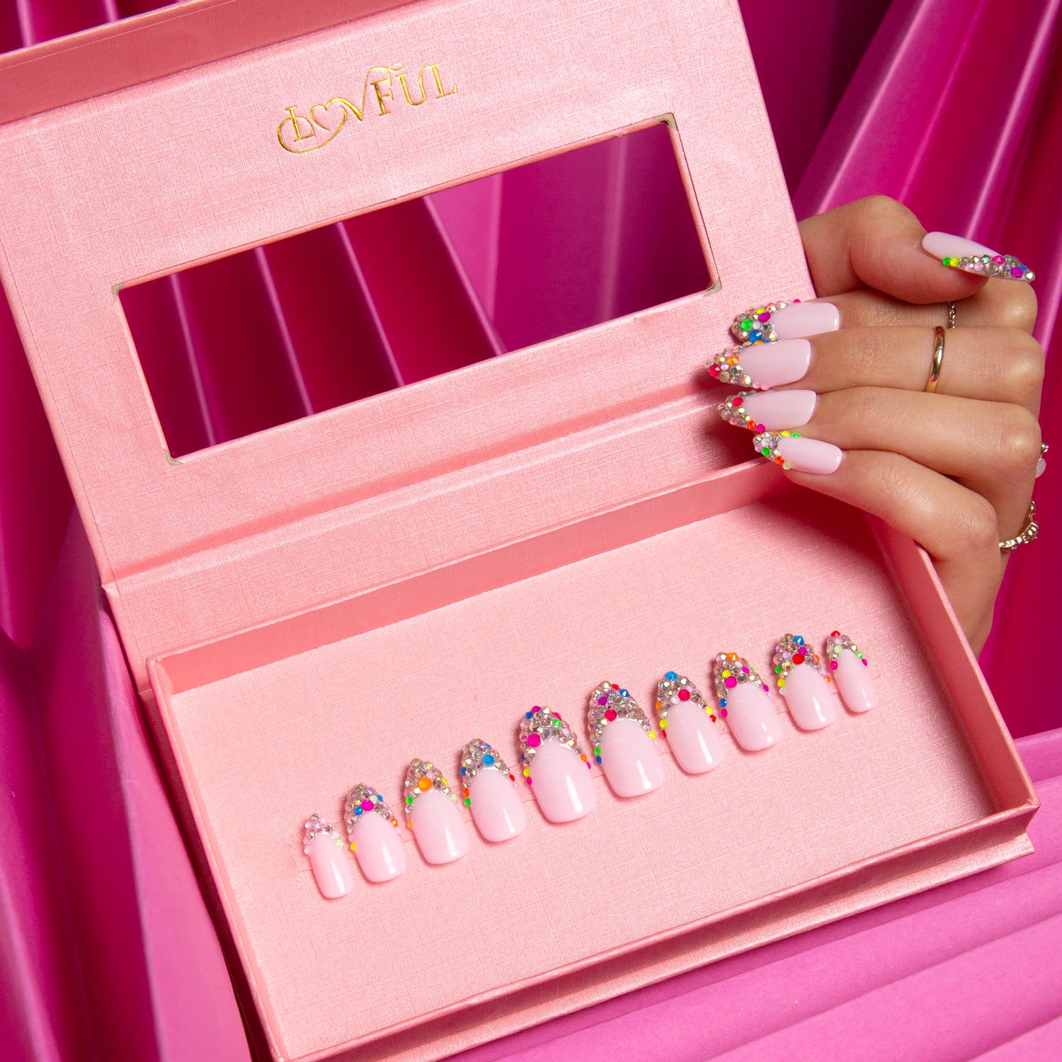 Hand holding pink box of Lovful 'Jelly Bean' press-on nails. The nails feature a chic nude base with colorful sugary beans on the tips. Vibrant pink background.