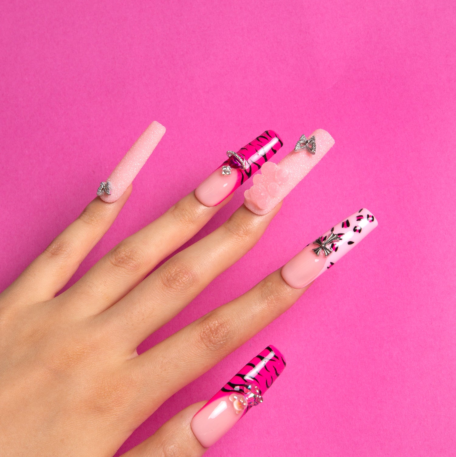 Hand with long square press-on nails with hot pink French tips, leopard prints, zebra patterns, glittery accents, and charms on vibrant pink background