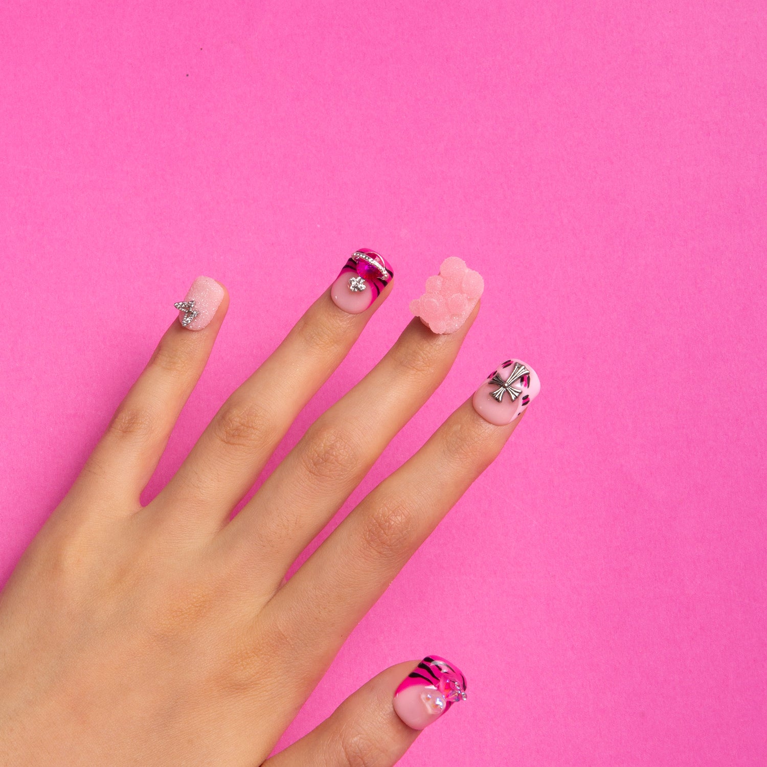 Hand with Safari Love press-on nails featuring hot pink French tips, leopard prints, lips, butterflies, and charming accents against a pink background.
