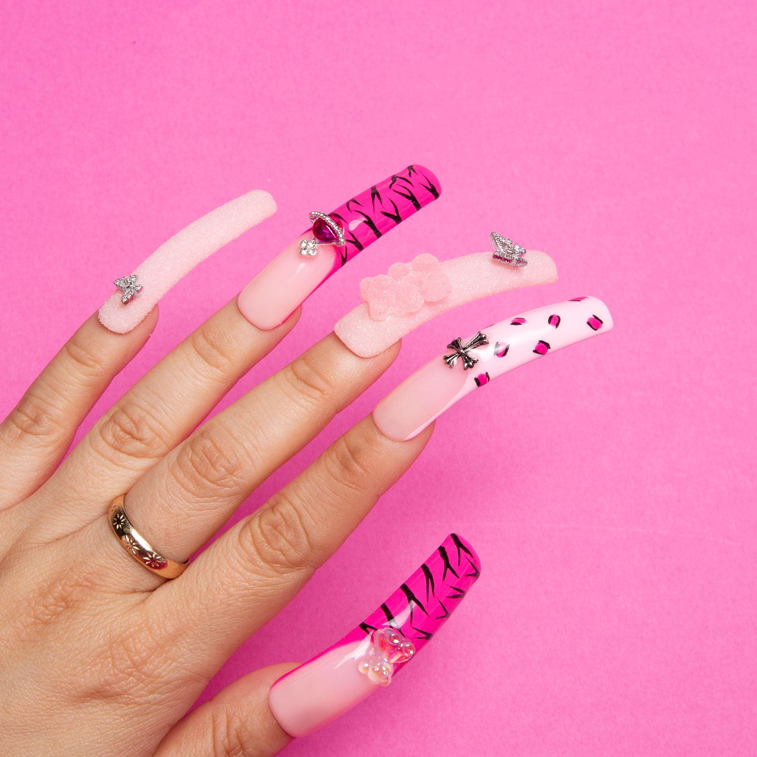 Close-up of hand with Safari Love Curve press-on acrylic nails featuring hot pink French tips with leopard prints, lips, butterflies, rhinestones, and a flower against a bright pink background.