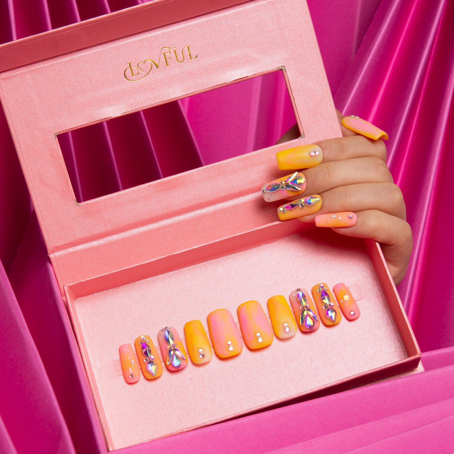 Lovful 'Sunset Slushy' press-on nails in orange and yellow tones with rhinestone accents, displayed in an open pink box with a window. Hand with matching nails also visible.