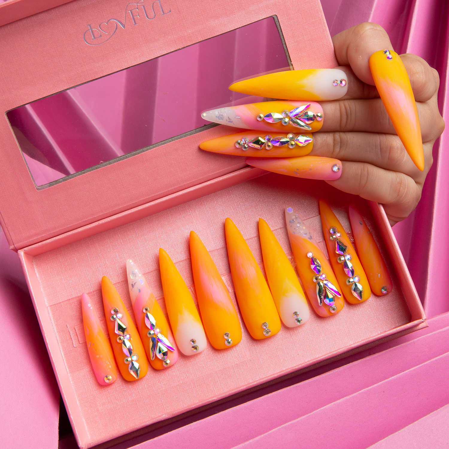 Lovful Sunset Slushy press-on stiletto nails in a pink box, featuring orange and yellow tones with radiant rhinestones, held by a hand.