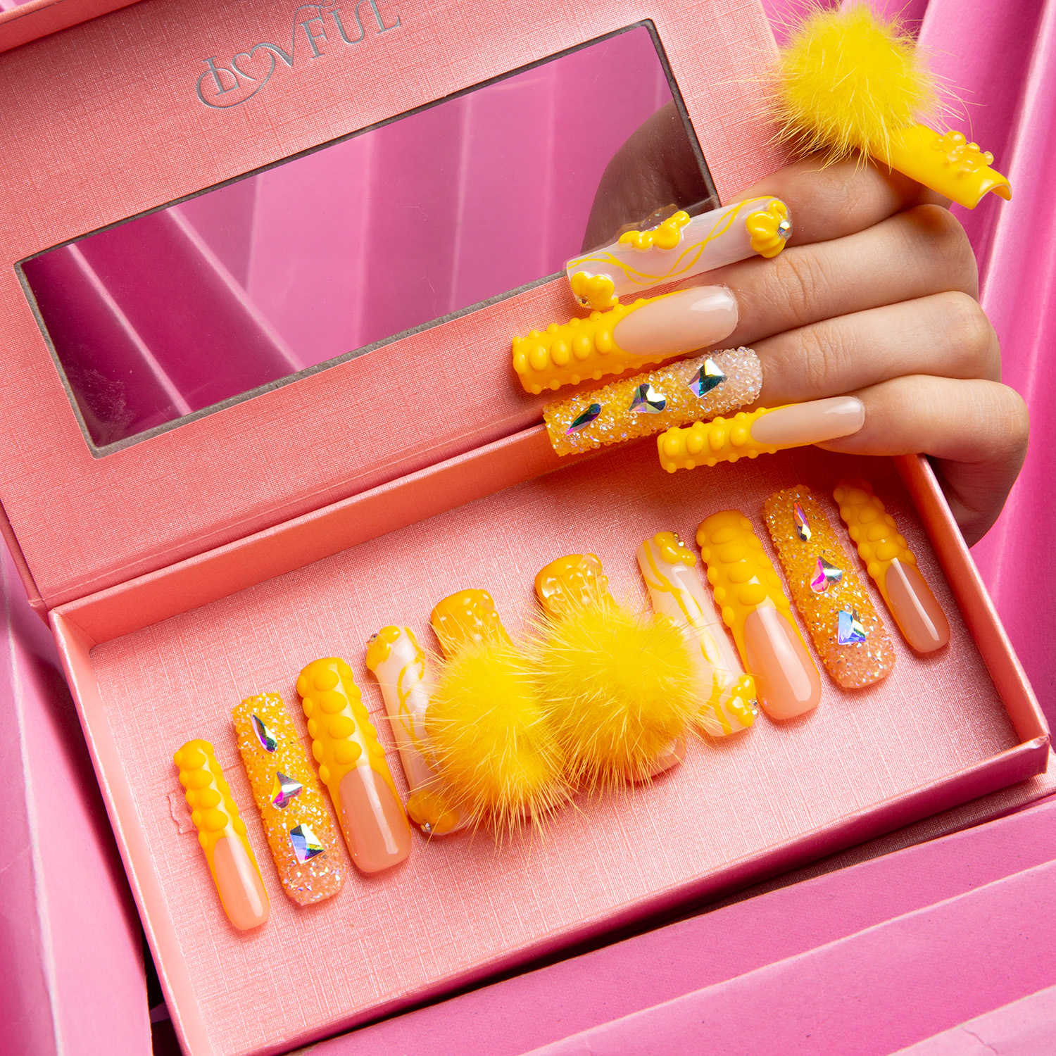 Hand with yellow-themed press-on acrylic nails adorned with glitter and gems, and fluffy ball decorations, placed in a pink Lovful box