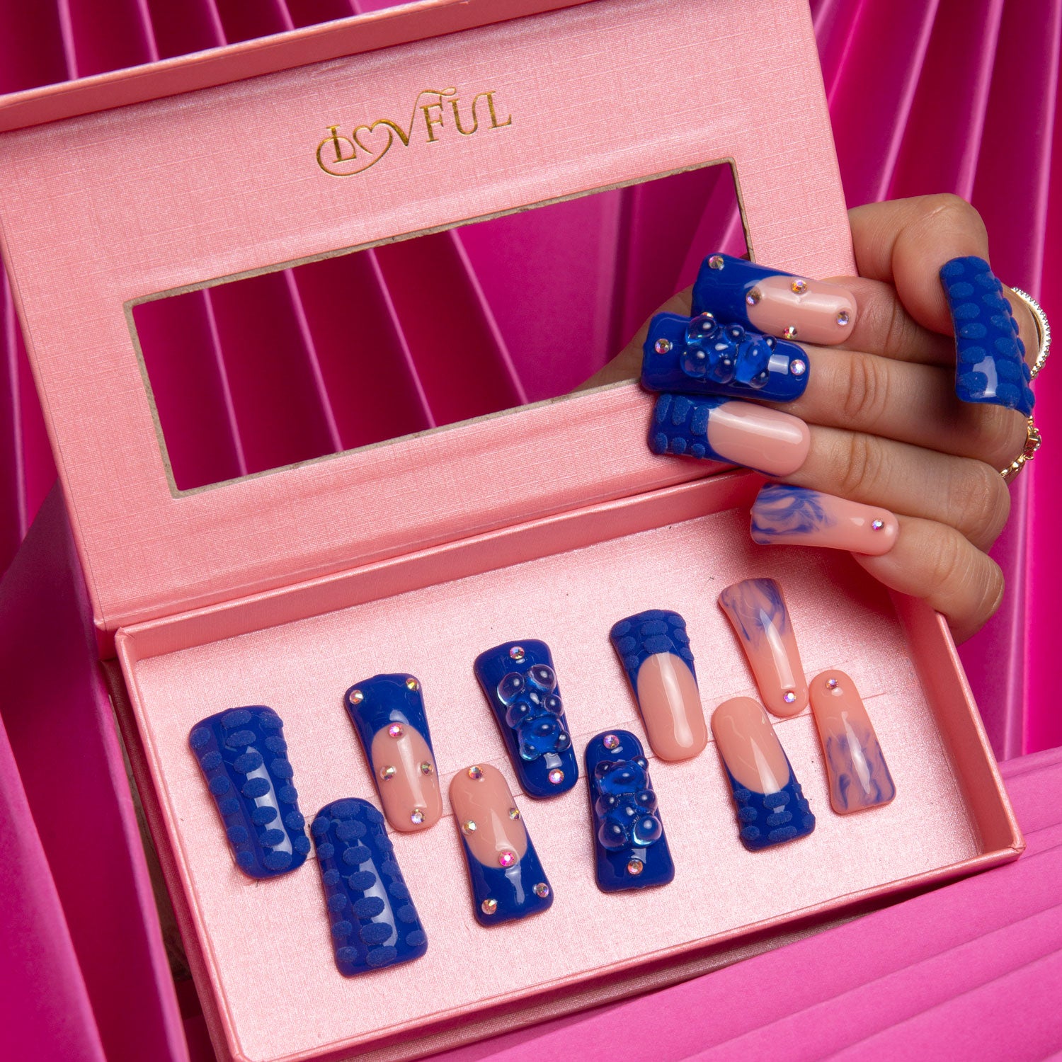 Lovful press-on nails set in a pink box with blue rhinestone gummy bear designs and hand-drawn 3D dots, displayed on a hand against a bright pink background