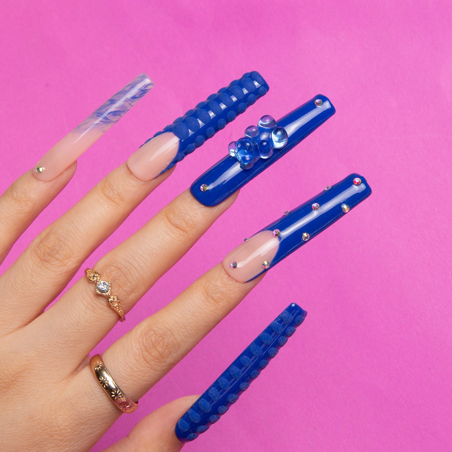 Hand with long square press-on nails in bright blue designs, adorned with French tips, rhinestones, and 3D gummy bear decorations, wearing gold and diamond rings.