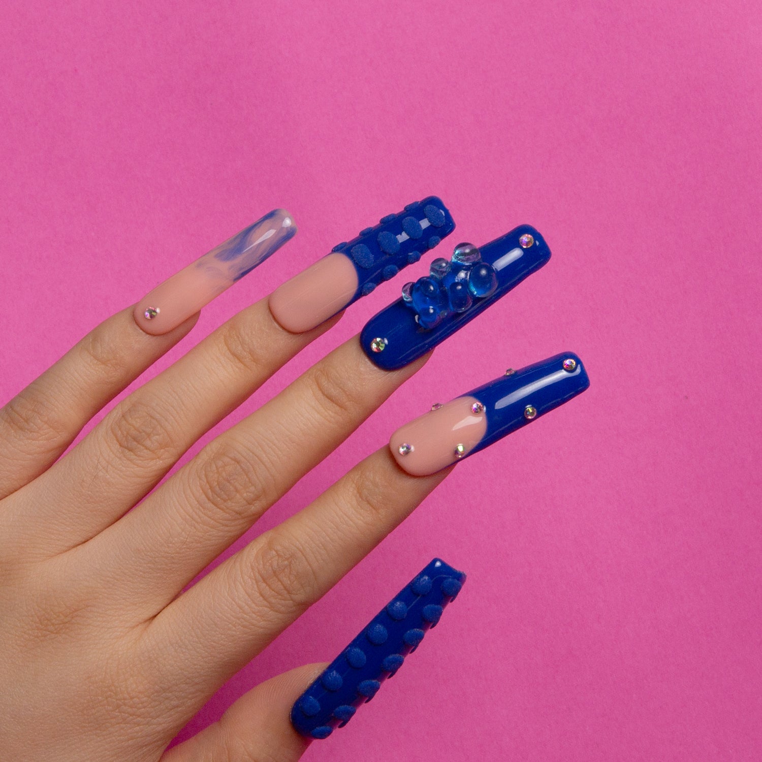 Blue and pink press-on acrylic nails with rhinestones, gummy bear decorations, and 3D dots on a hand against a pink background. Square nail shape from Lovful.com's Little Blue Bear collection.