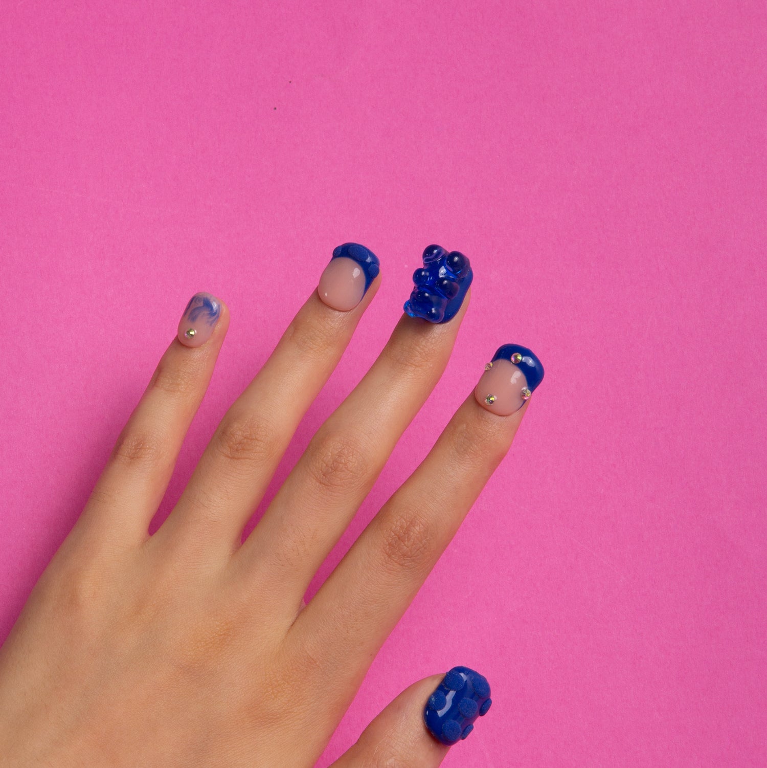 Hand sporting 'Little Blue Bear' square-shaped press-on French tip nails with blue hue, rhinestones and 3D gummy bears on a pink background.