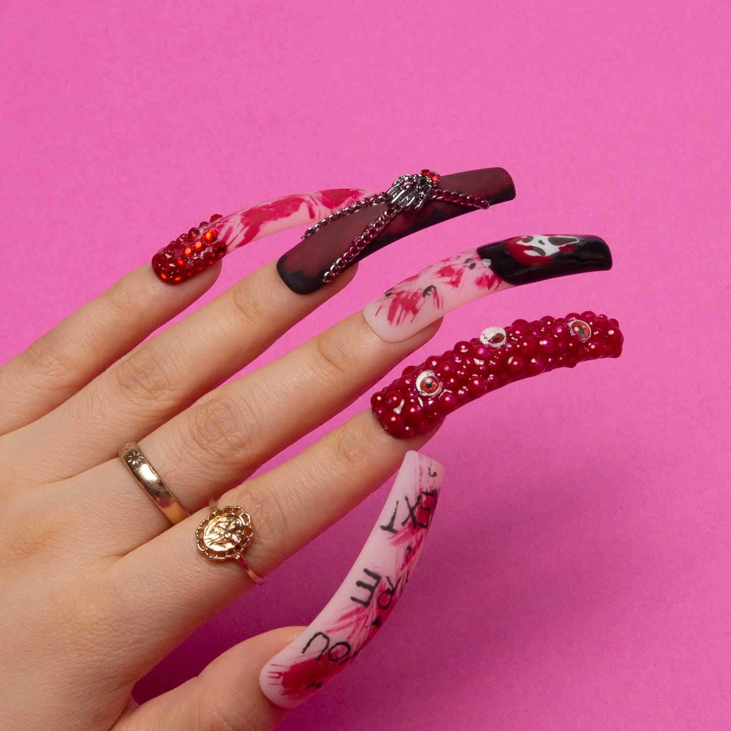 Hand with 'You Are Next' press-on nail set featuring ghostface design, eerie eyes, red gems, and blood-red splashes on a bright pink background for Halloween manicure.