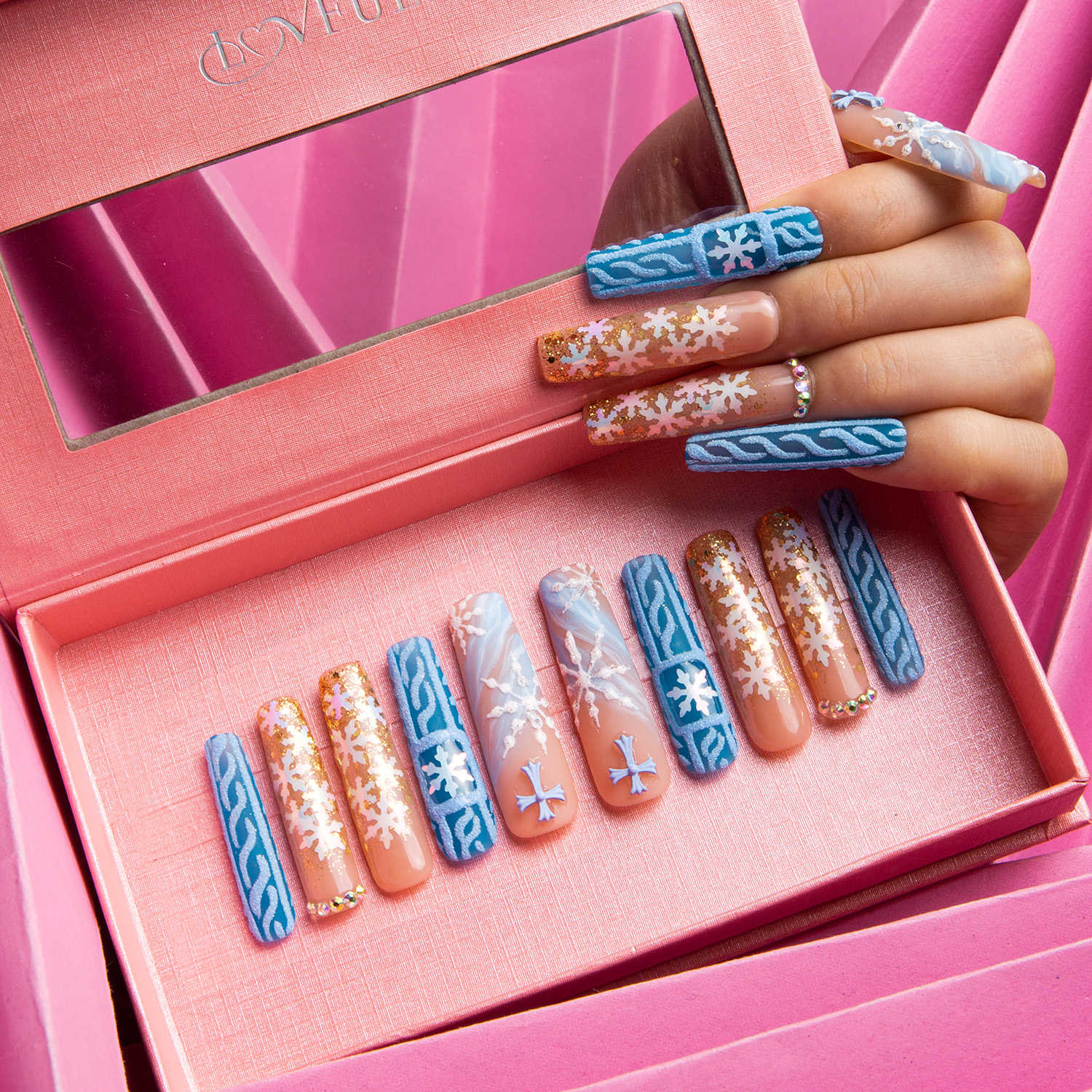 Winter Sunshine press-on nails with blue, gold glitter, and white snowflake designs in a pink box with a mirror lid, held by a hand with matching nails.