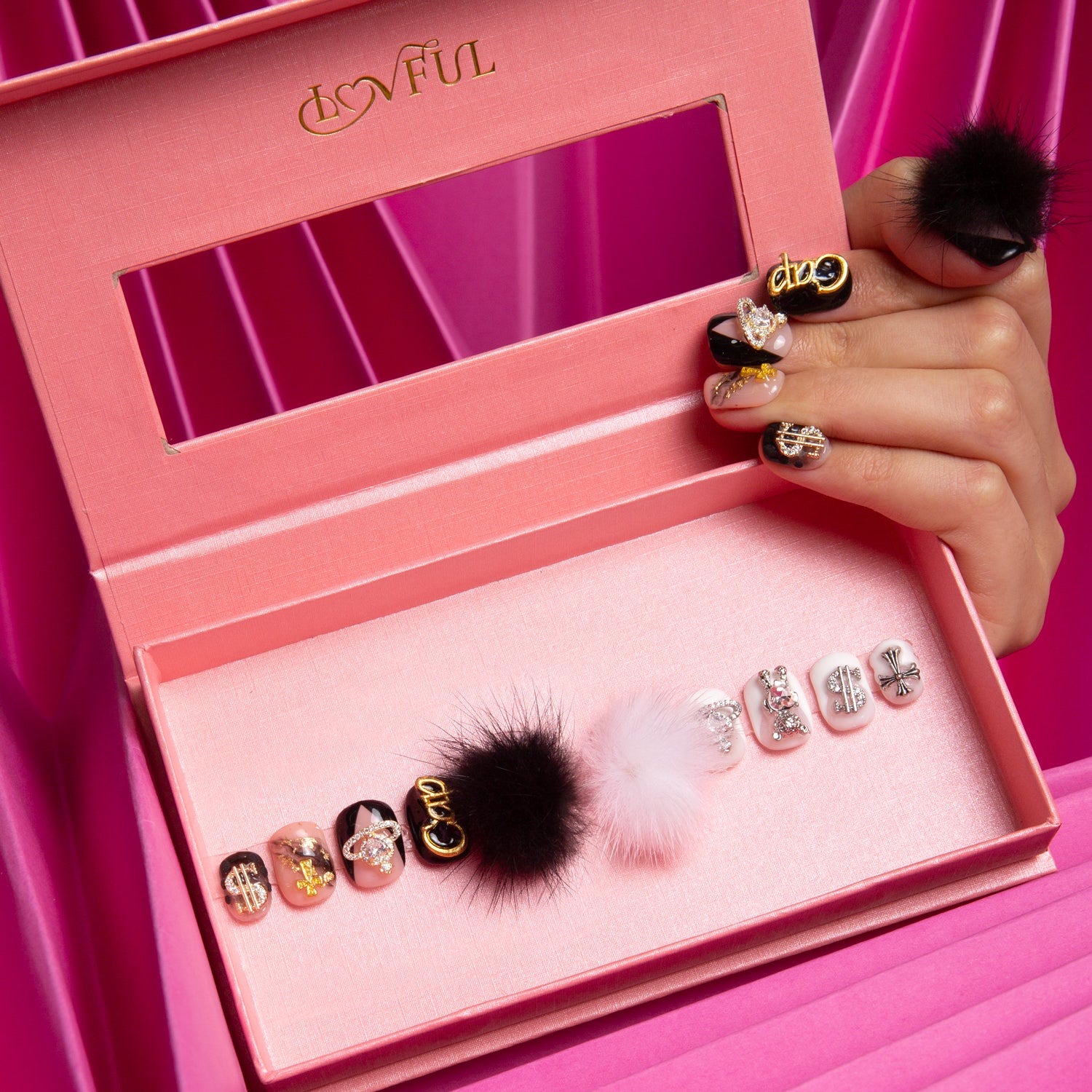 Pink box displaying a set of Capricorn-themed press-on nails with detachable black and white fluffy balls and small intricate decorations, held by a hand with matching nails