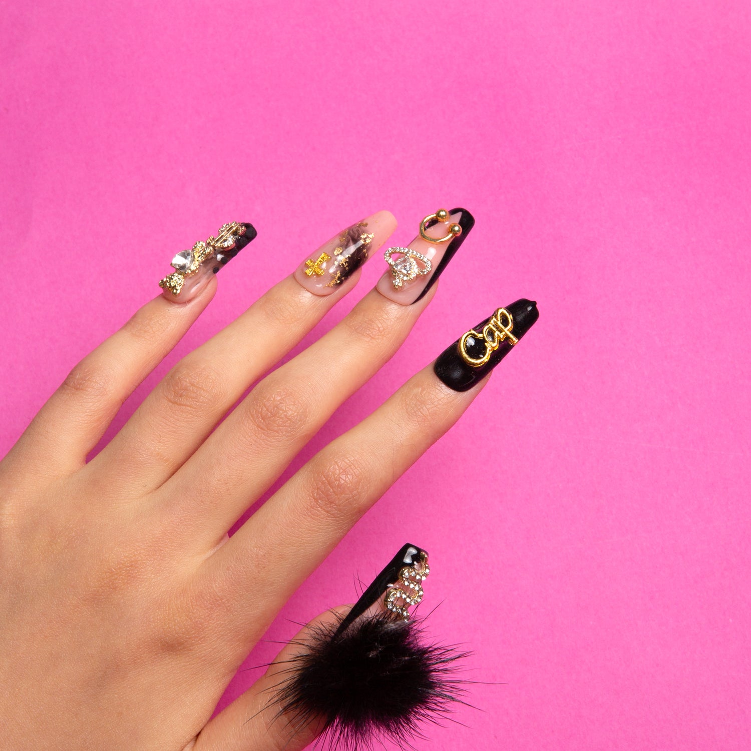 Hand with Capricorn-inspired press-on nails featuring black and white color palette, gold decorations, gemstones, and black fluffy balls on a pink background