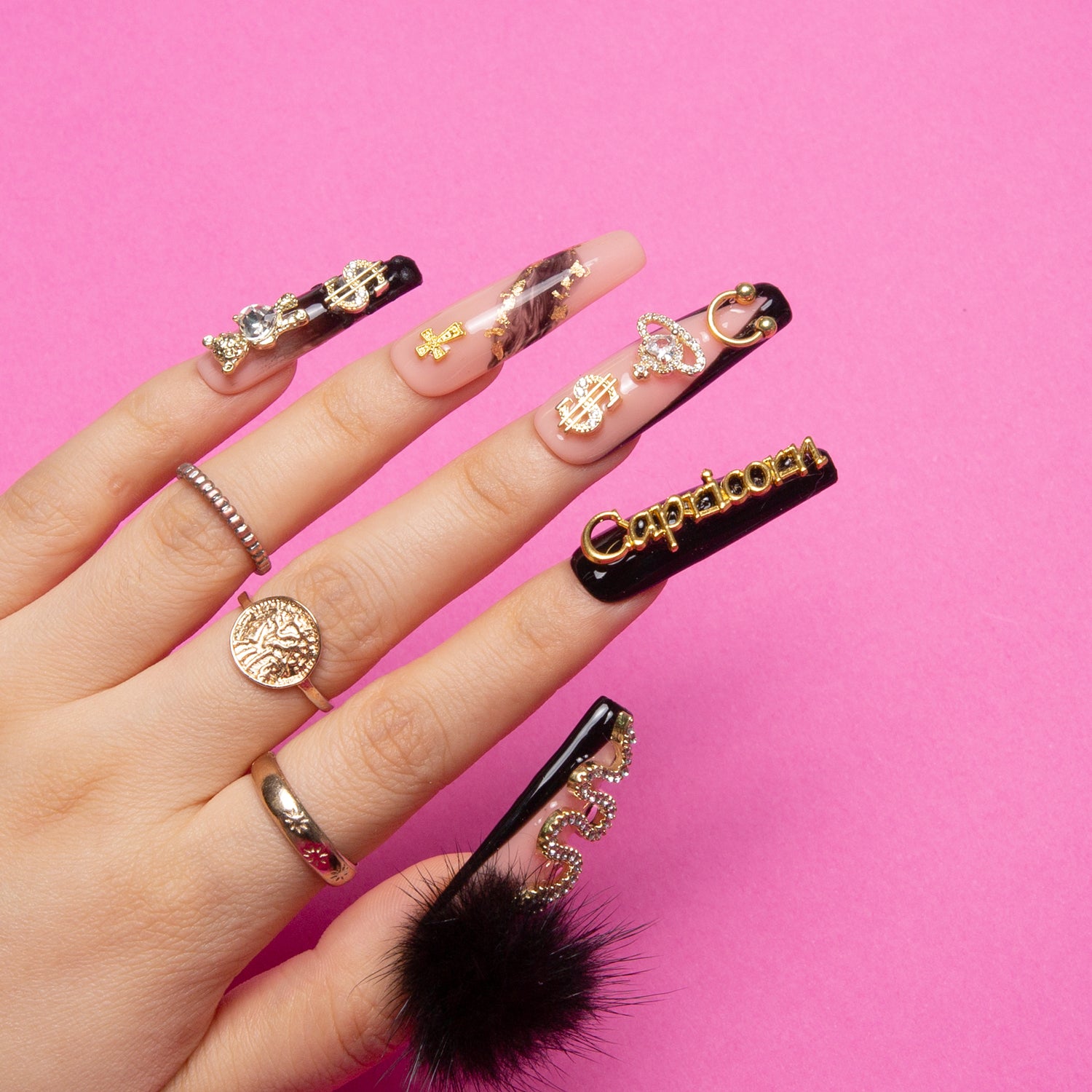 Hand showcasing Capricorn-themed press-on nails with gold decorations and detachable black fluffy balls on black and white nails against a pink background. Nails embody Capricorn's disciplined yet adventurous spirit.