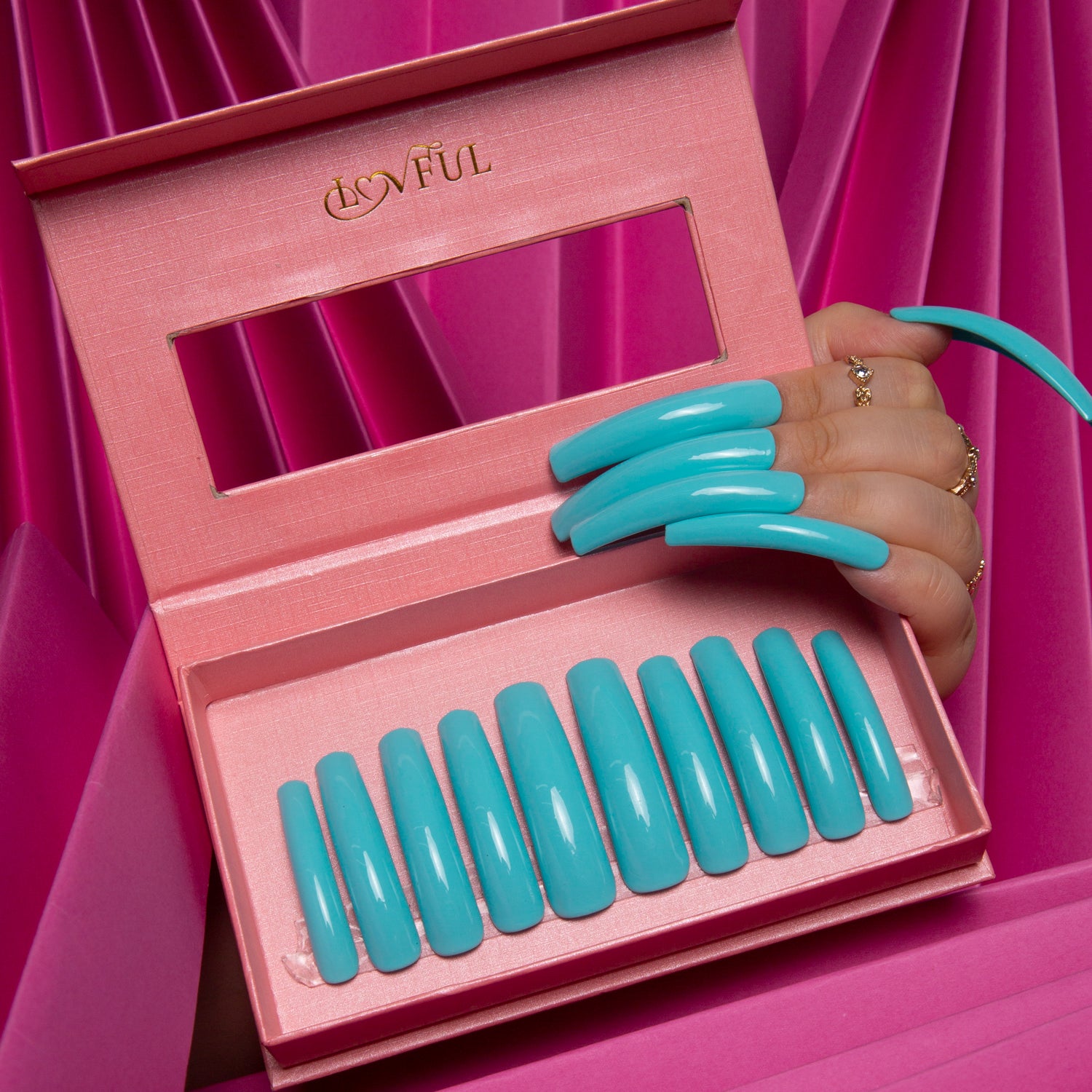 Shiny light blue press-on acrylic nails displayed in a pink Lovful box, held by a hand with matching nails in front of a pink pleated fabric background.