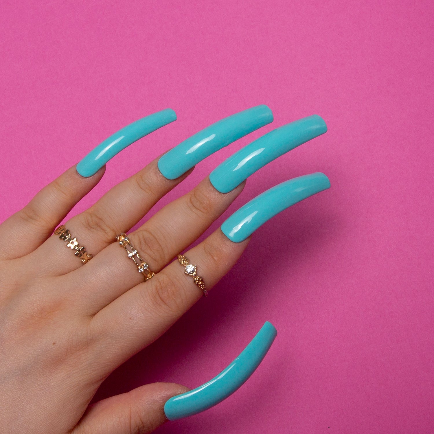 Hand showcasing long, curved shiny blue press-on nails against a pink background, adorned with multiple gold rings. Perfect for embracing stylish vacation vibes and beach-inspired looks.