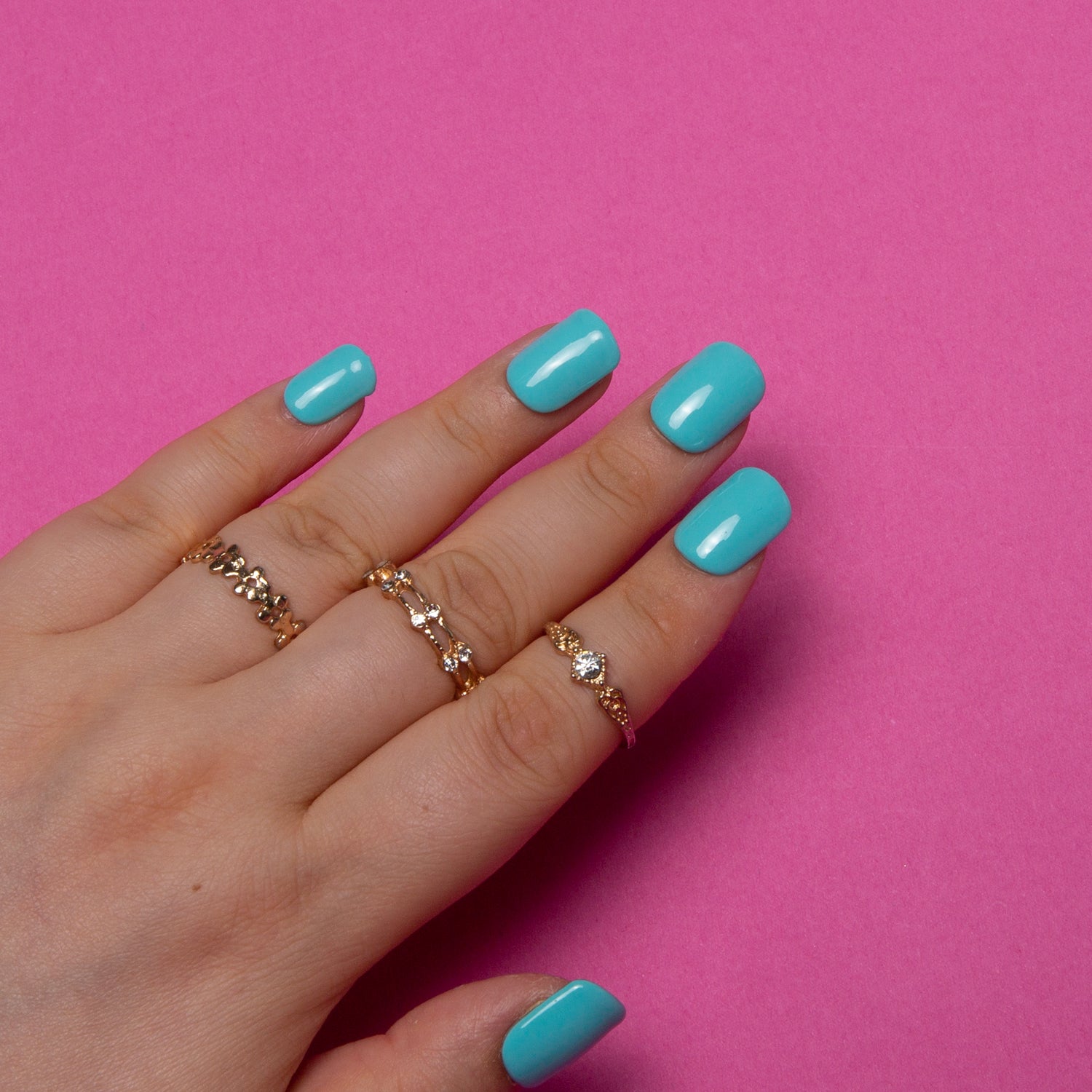 Hand with shiny blue square-shaped press-on nails from Lovful.com against a pink background. The hand is wearing gold rings with small gemstones, showcasing the 'Shiny Blue' nail design which captures the essence of seaside getaway and beach vibes.