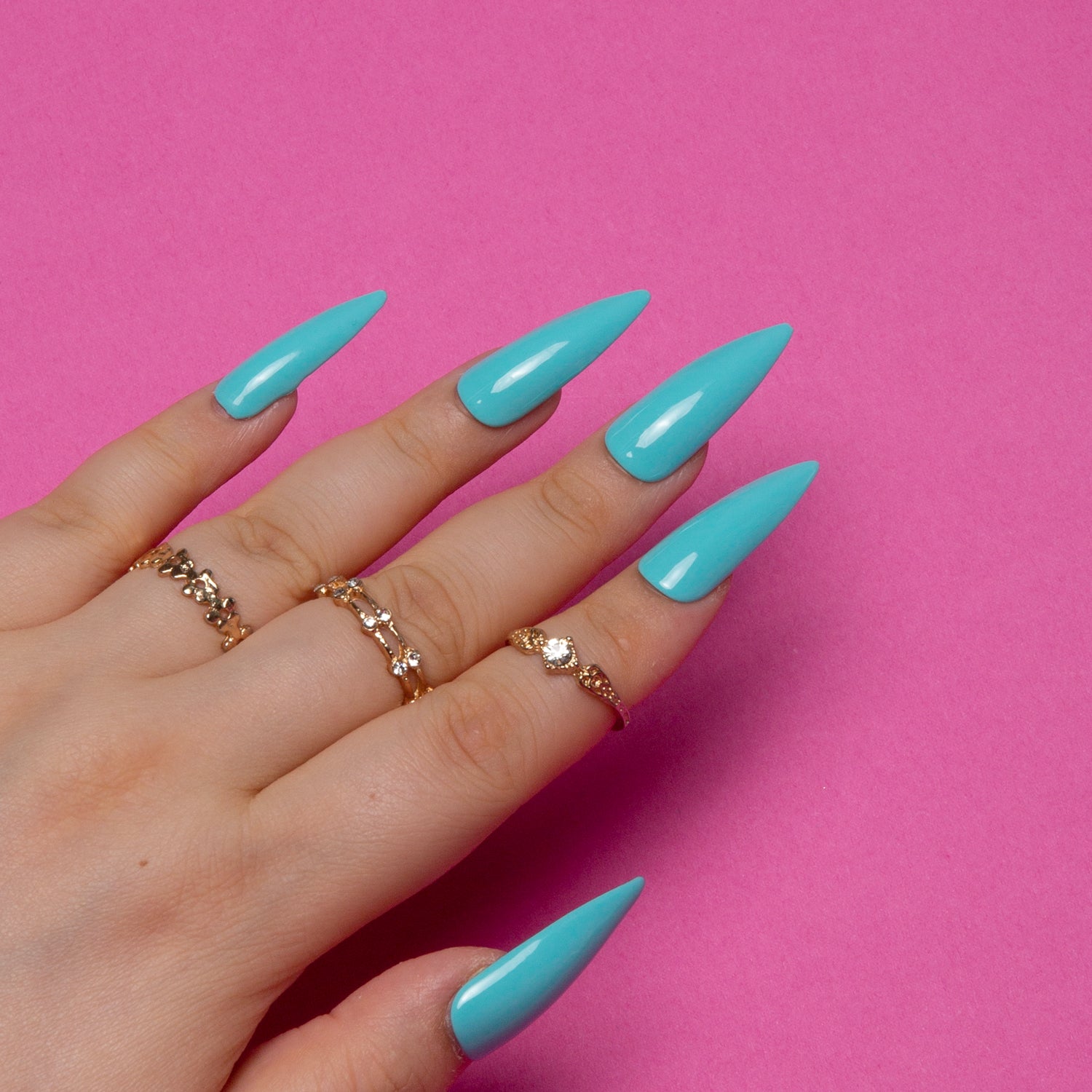 Hand with shiny light blue stiletto press-on nails adorned with delicate gold rings against a pink background.