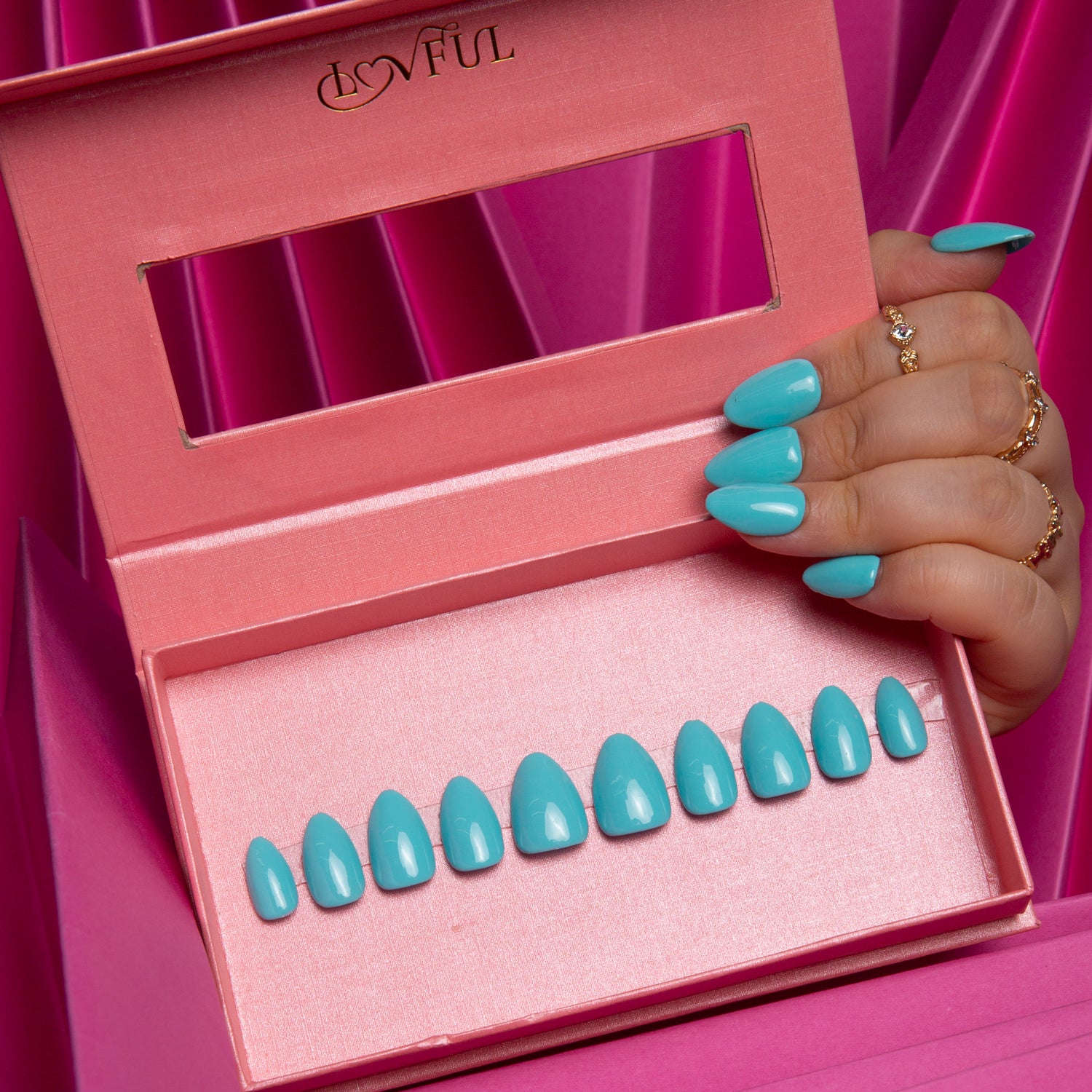 Hand displaying shiny blue almond-shaped press-on nails over an open pink box containing a set of similar blue nails with the 'Lovful' brand name on the lid. Background of magenta draped fabric.