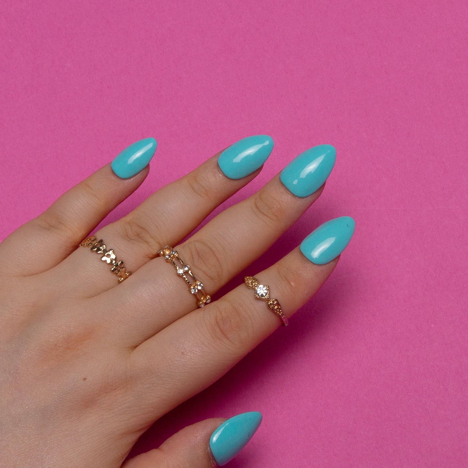 Hand with almond-shaped shiny blue press-on nails and gold rings against a pink background, showcasing the Shiny Blue nails from Lovful.com.