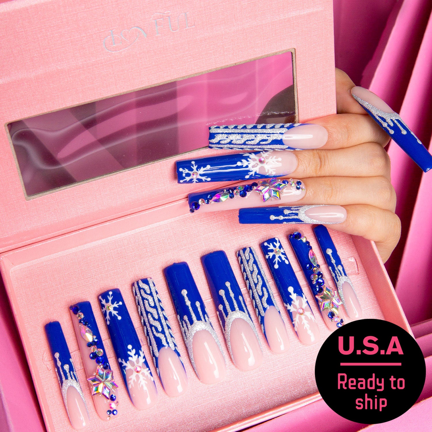 Close-up of a hand with blue press-on nails featuring white snowflake designs and colorful gemstones, arranged in a pink box with 'U.S.A Ready to ship' text.