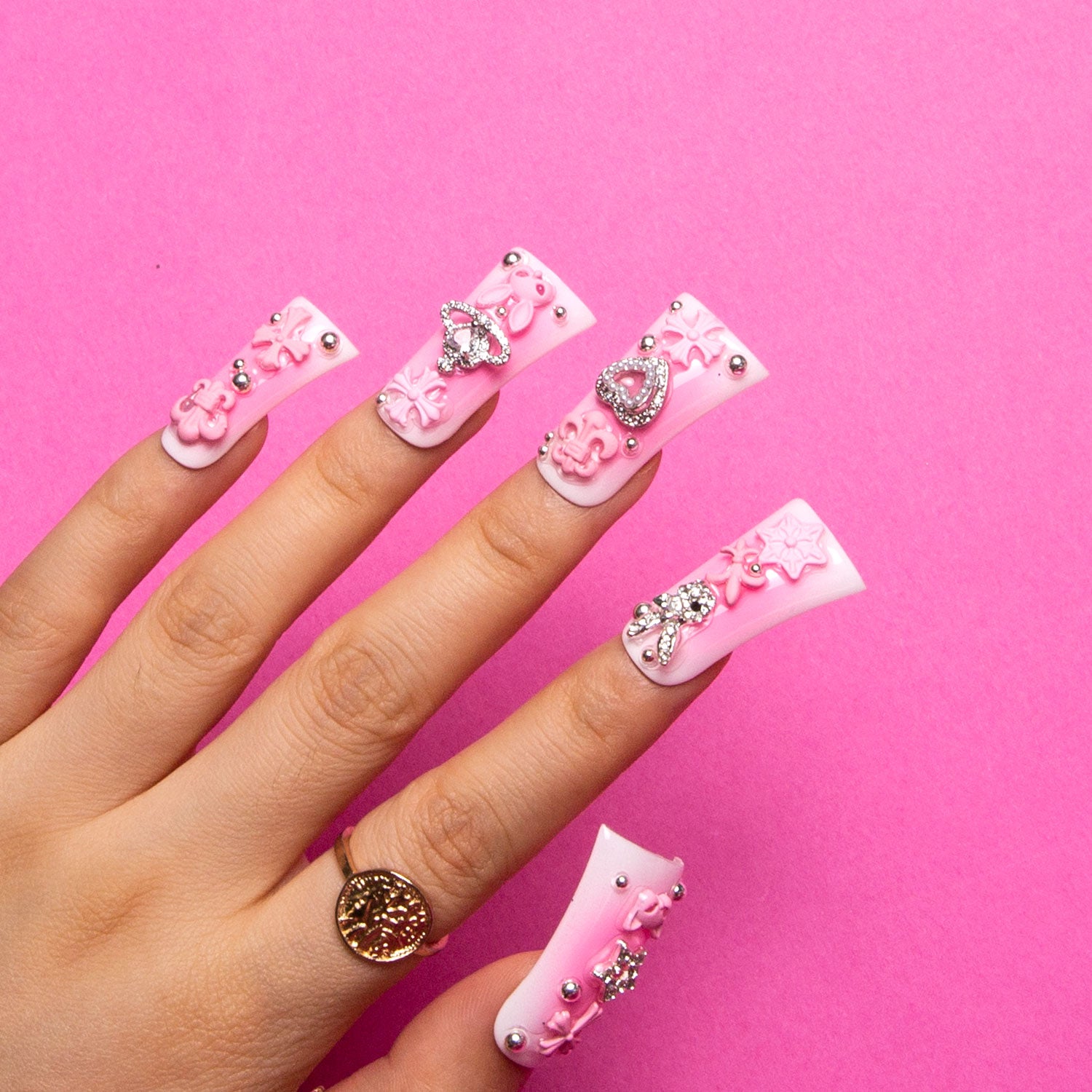 Hand with 'Pink Bliss Blush' gradient press-on acrylic nails by Lovful, adorned with pink crosses, cat designs, heart shapes, planet motifs, rhinestones, and pearls on duck-shaped nails.