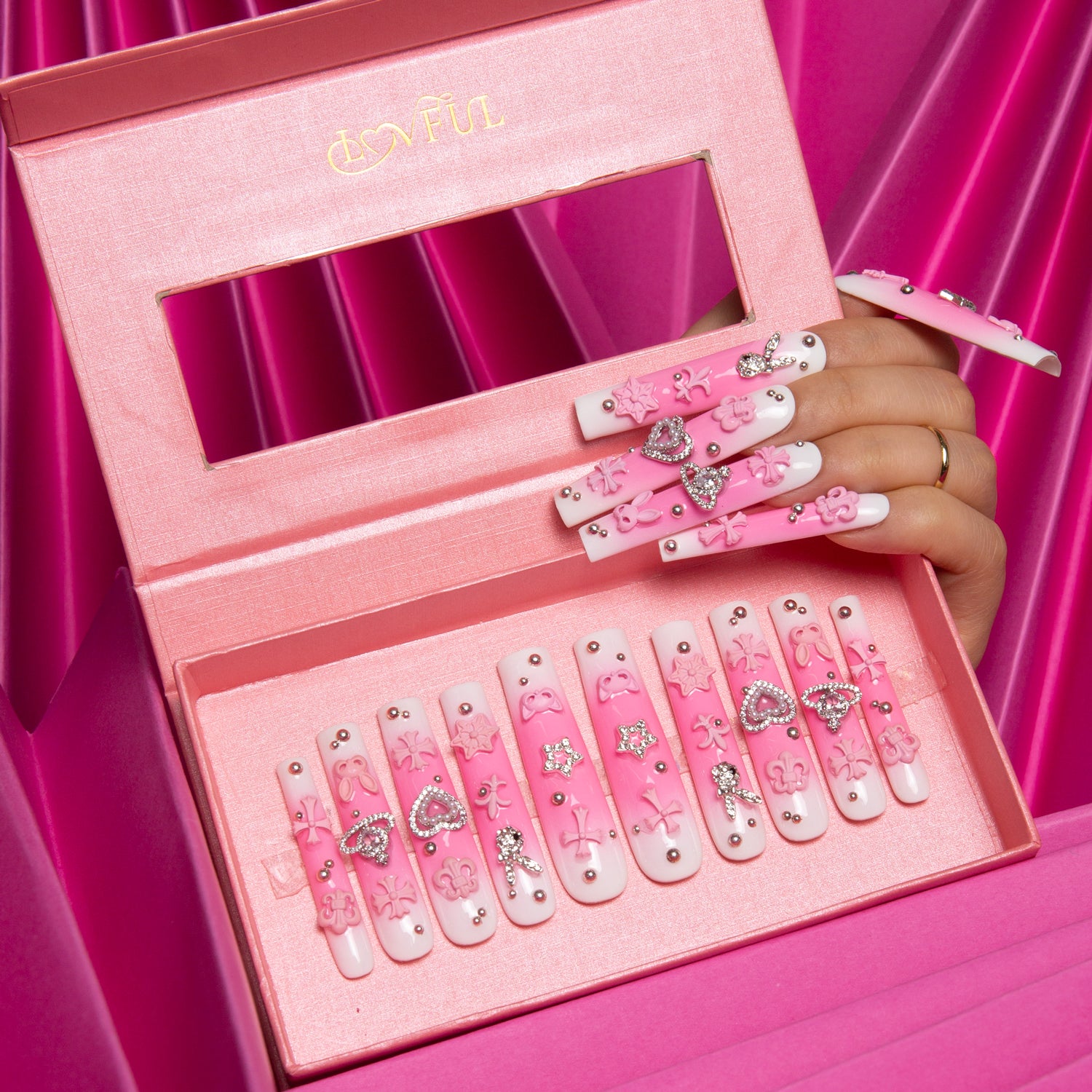 Press-on nails from Lovful's 'Pink Bliss Blush' collection in a pink box. Nails have a pink gradient style with heart shapes, pink crosses, cat designs, and planet motifs. A hand models the nails.