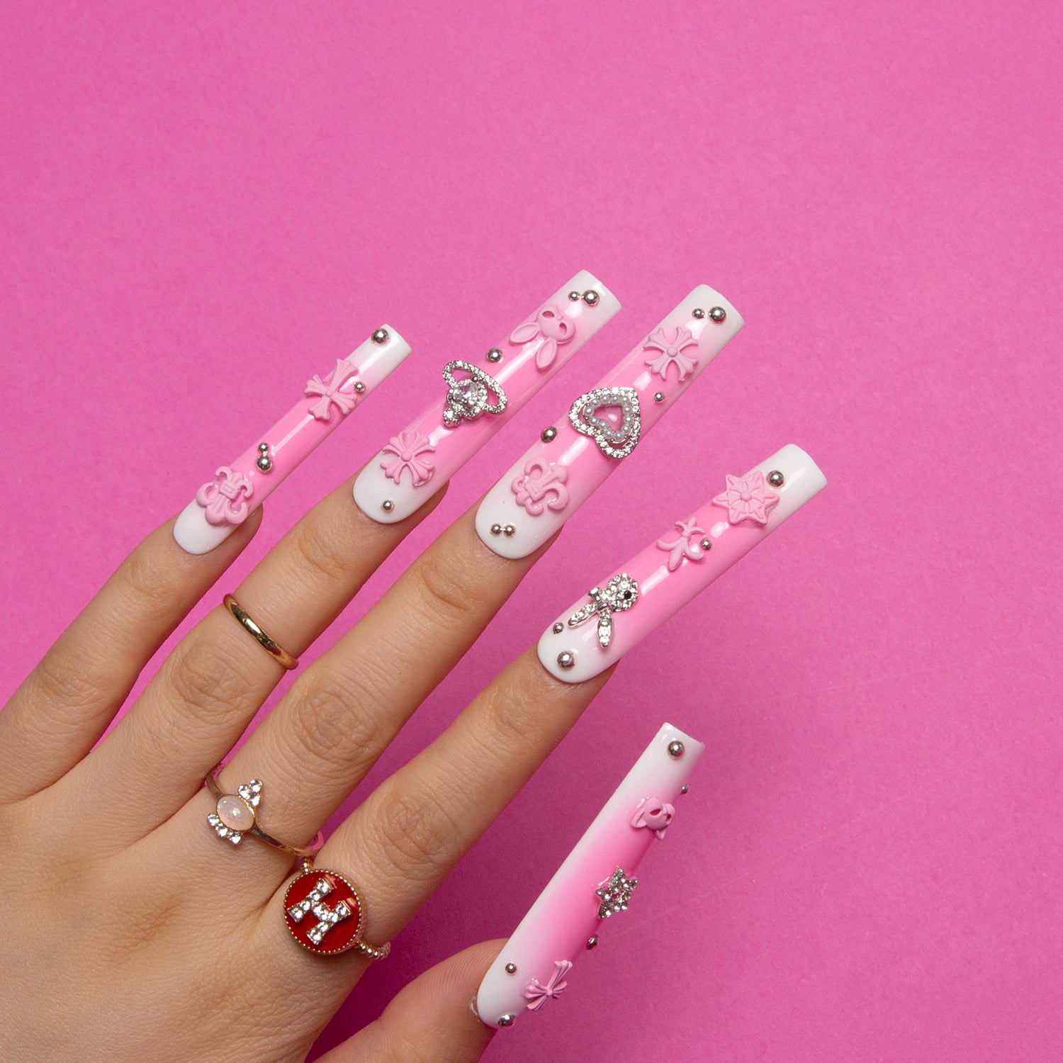 Hand with square gradient pink blush press-on nails adorned with crosses, cat designs, hearts, and planets on a pink background, showcasing whimsical and romantic nail art.