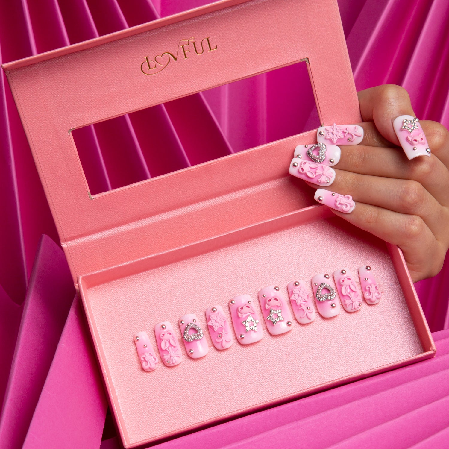 Set of 'Pink Bliss Blush' press-on nails by LOVFUL featuring gradient pink shades with cute and cosmic decorations, displayed in a branded pink box and applied on a hand against a pink fabric background.