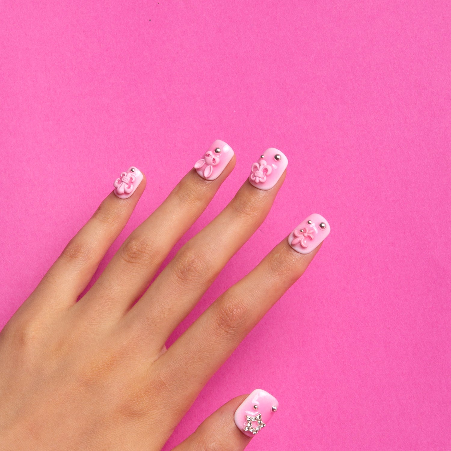 Hand with Pink Bliss Blush square press-on nails featuring gradient pink blush effect and adorable decorations like pink crosses, whimsical cat designs, heart shapes, and planet motifs.