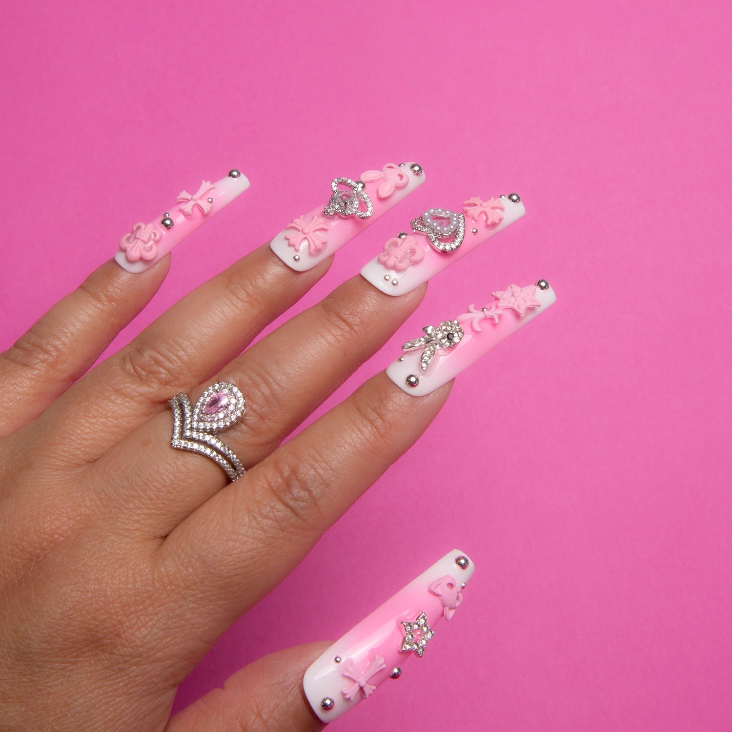 Coffin-shaped press-on nails set named 'Pink Bliss Blush,' featuring pink blush gradient with cute pink crosses, whimsical cat shapes, hearts, and celestial star and planet motifs, worn on a hand against a pink background.