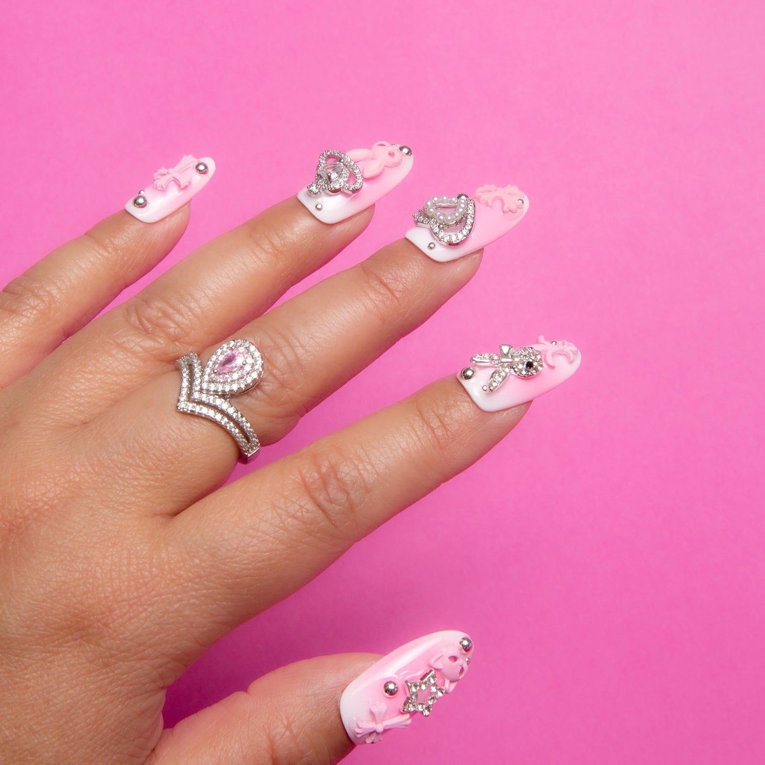 Hand displaying Pink Bliss Blush press-on nails with gradient pink blush effect and adorable decorations including pink crosses, cat designs, hearts, and planets. Decorative ring with pink gemstone on the ring finger.