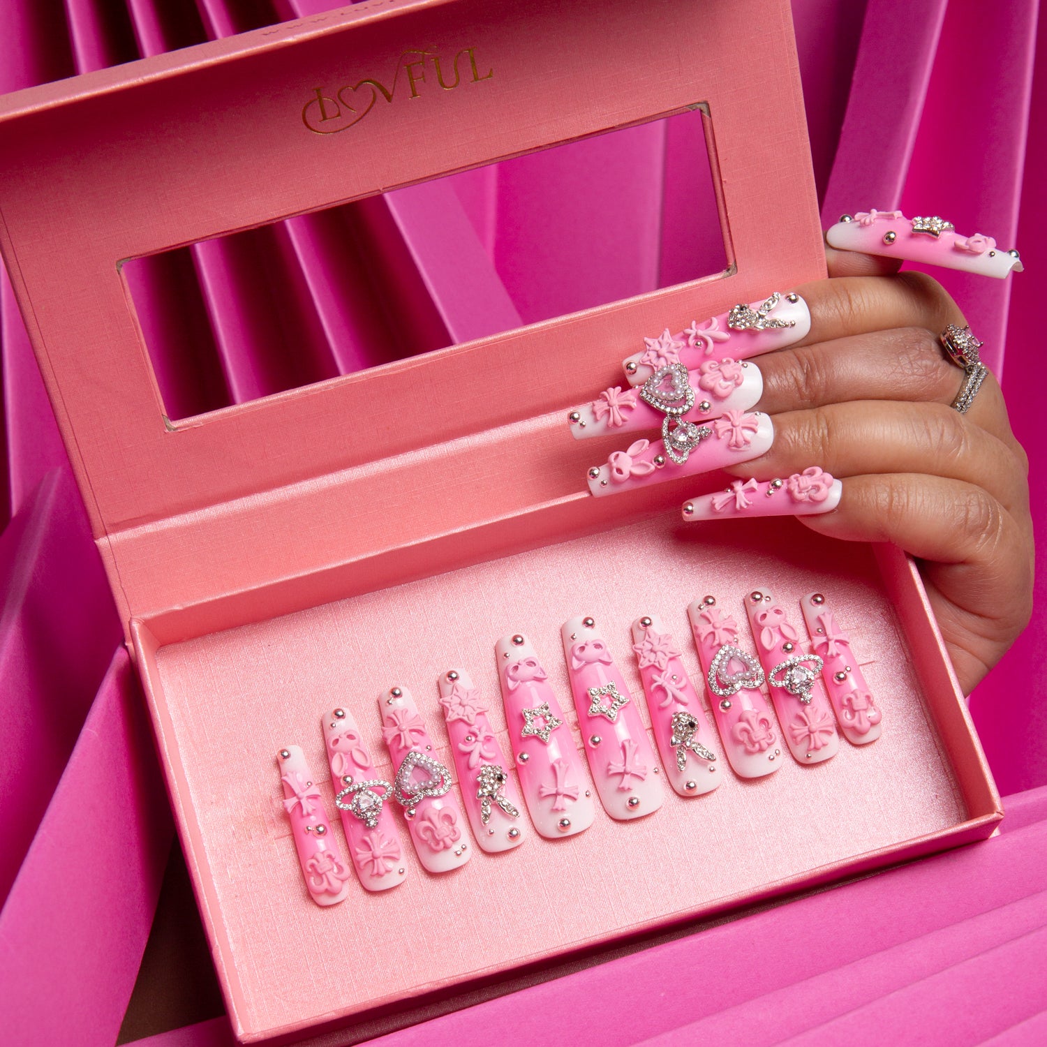 Hand displaying decorated 'Pink Bliss Blush' press-on nails in a pink velvet box with gradient design, cute pink crosses, whimsical cat designs, heart shapes, and planet motifs. Lovful branding on the box, pink draped fabric background.