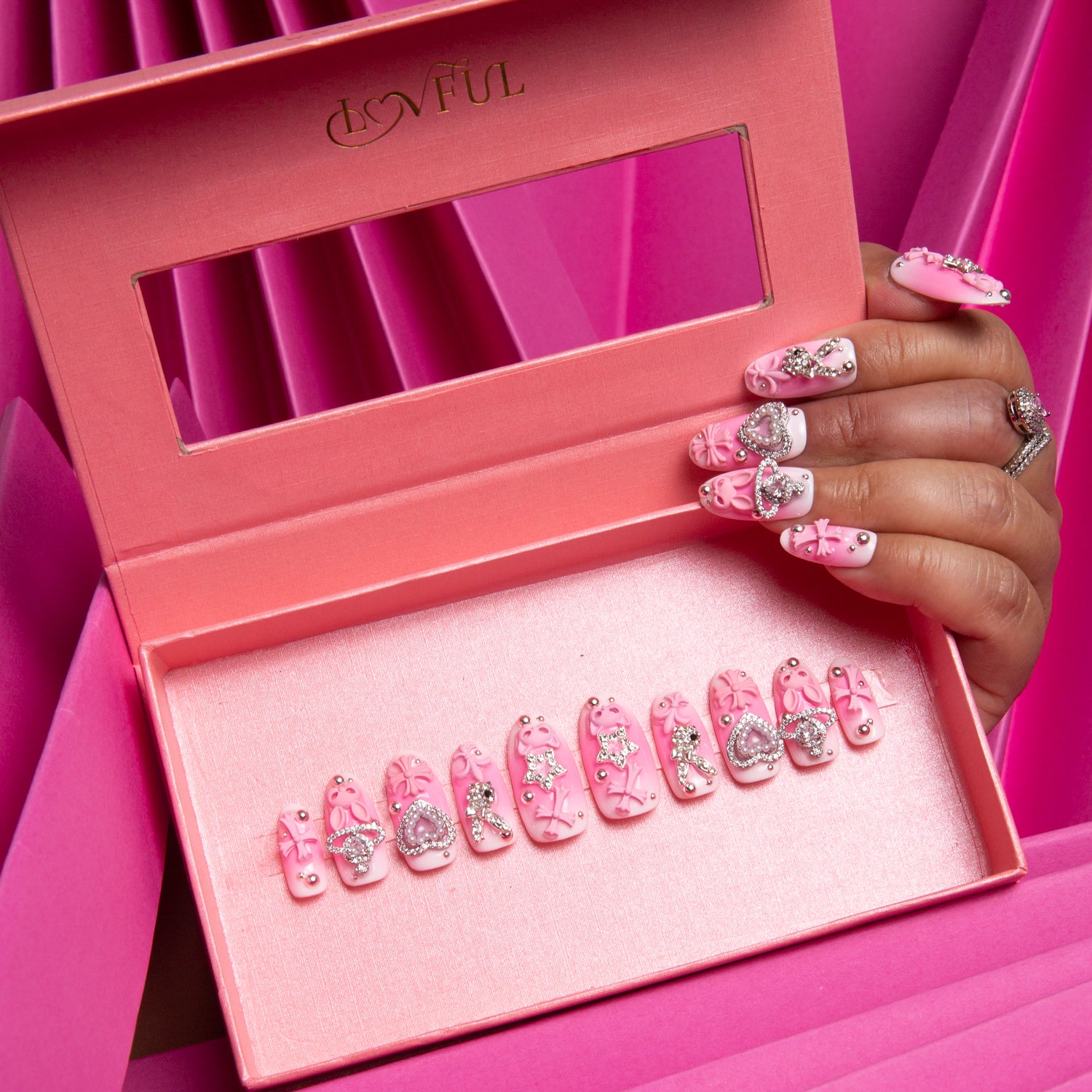 Lovful Pink Bliss Blush press-on nails set in a gradient pink blush style with decorations including cute crosses, cat designs, hearts, and planets. Hand with matching designed nails holding the box.