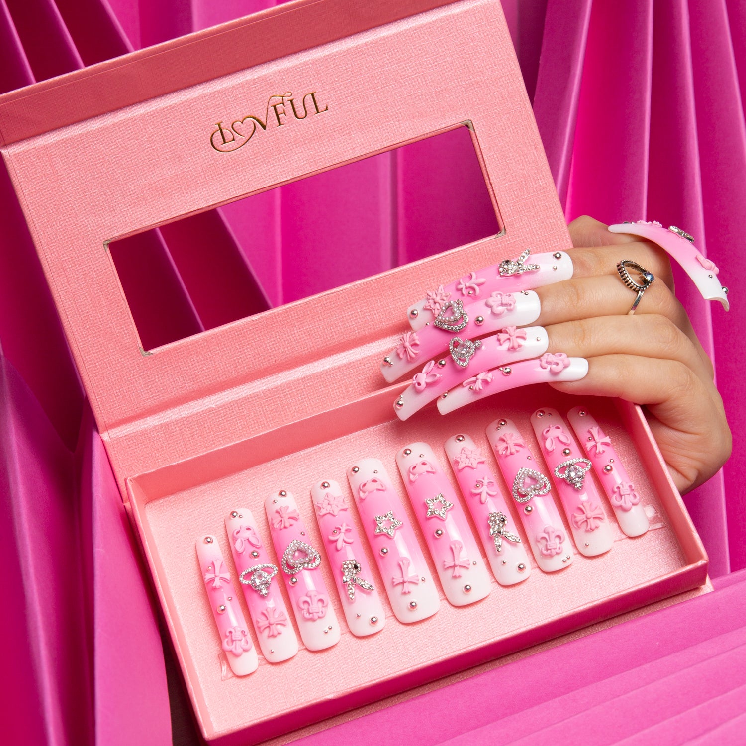 Pink Bliss Blush press-on nails in pink box by Lovful.com. Nails feature pink gradient with decorations including crosses, cats, hearts, and planets. Hand holding box against pink background.