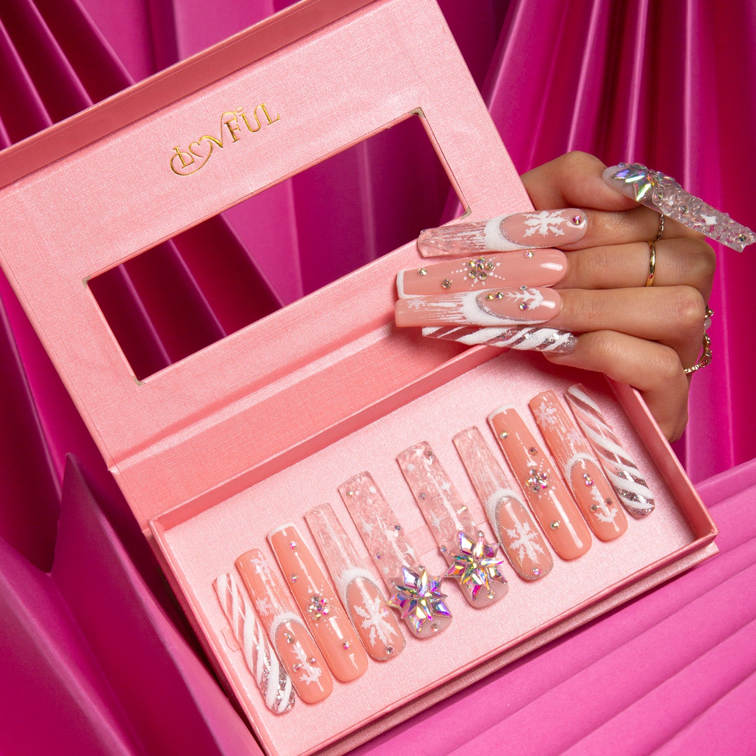 Hand displaying Lovful's Snowy Moonlight press-on nails with winter-themed designs including snowflakes and sparkles, neatly arranged in a pink box against a pink fabric background.
