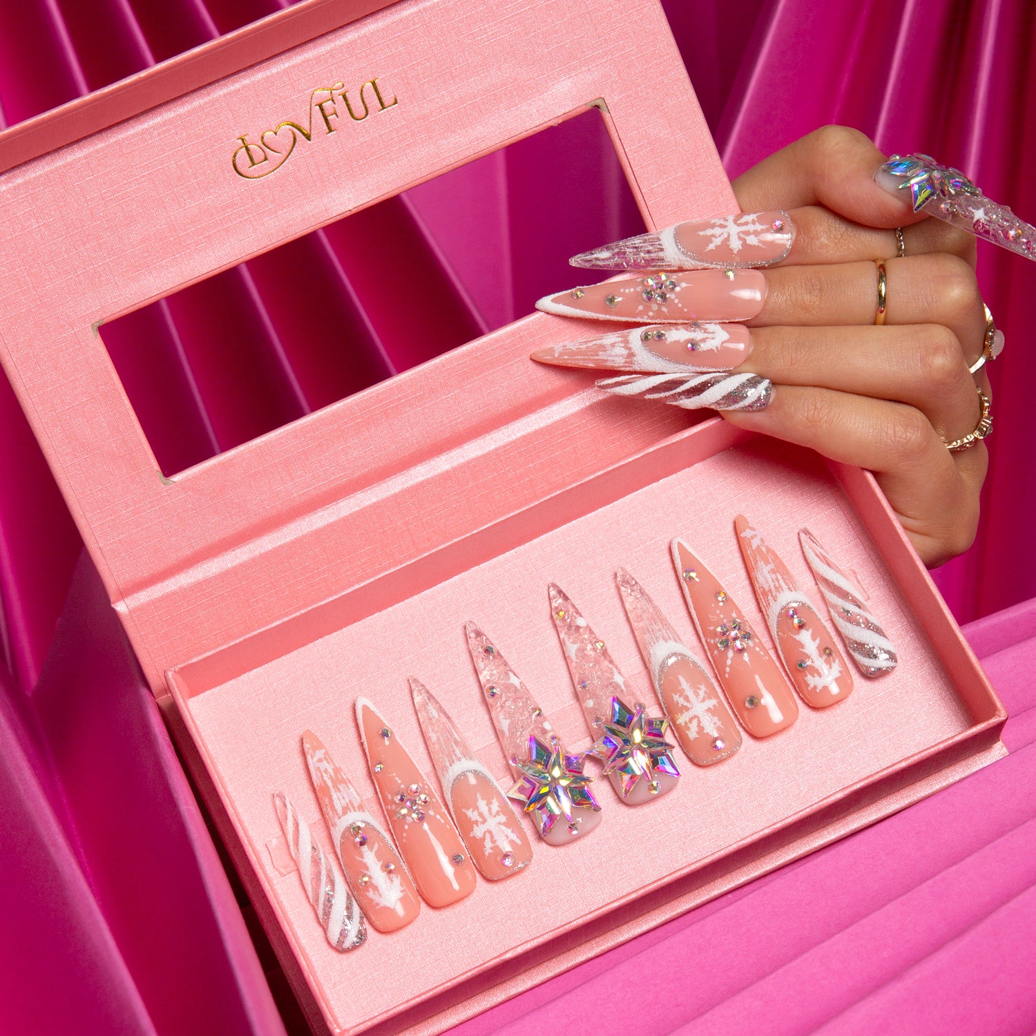 Lovful Snowy Moonlight stiletto press-on nails in pink box with snowflake designs and glitter, hand holding the box against pink satin background