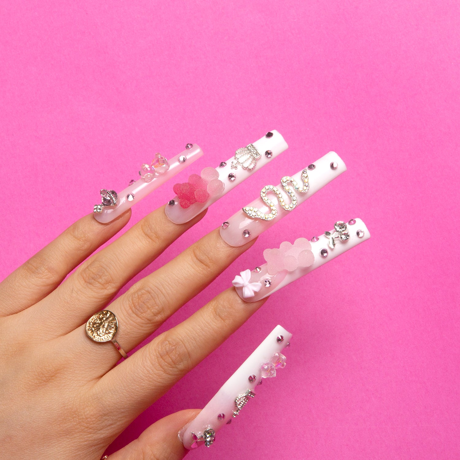 Hand with decorated press-on nails featuring pink gummy bears, rhinestones, floral designs, and a swan-shaped accessory against a pink background