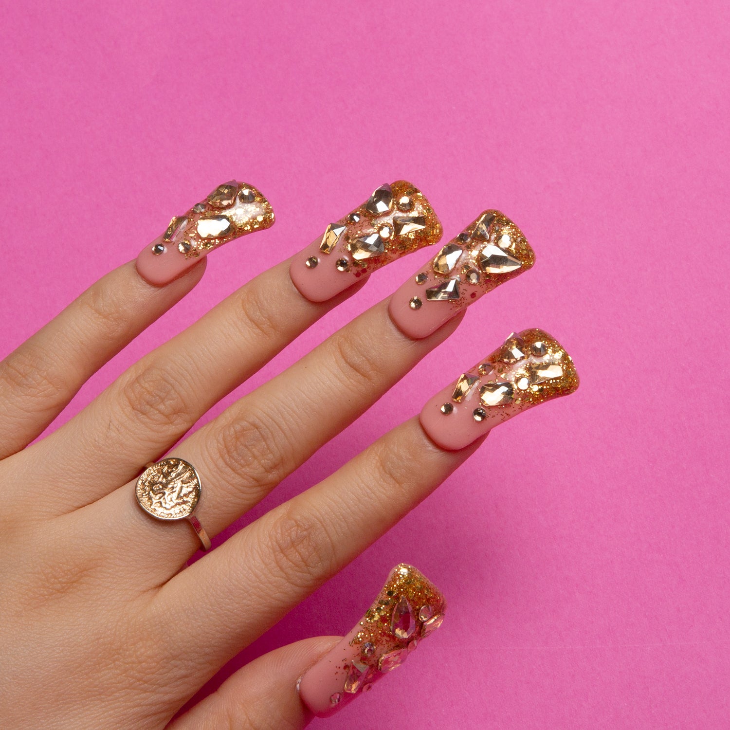 Hand with 24 pieces of press-on nails from Lovful's Stardust (H84) collection on a pink background. Nails feature golden French tips with glitter and jewel-like accents, emulating the elegance of a royal sunrise.