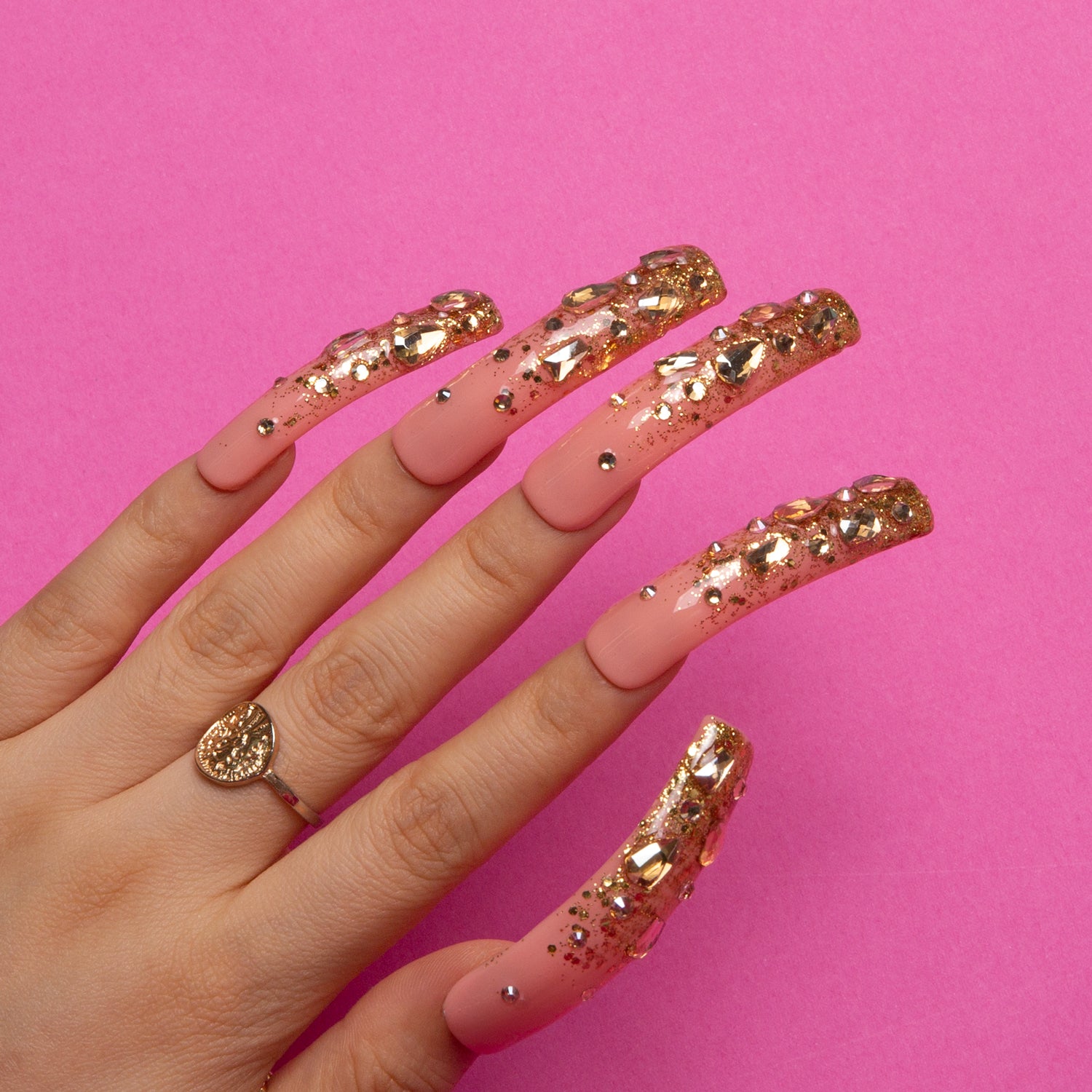 Hand with long press-on nails in the Stardust (H84) design, featuring pink bases and glittering golden French tips with sequins and rhinestones, against a pink background.