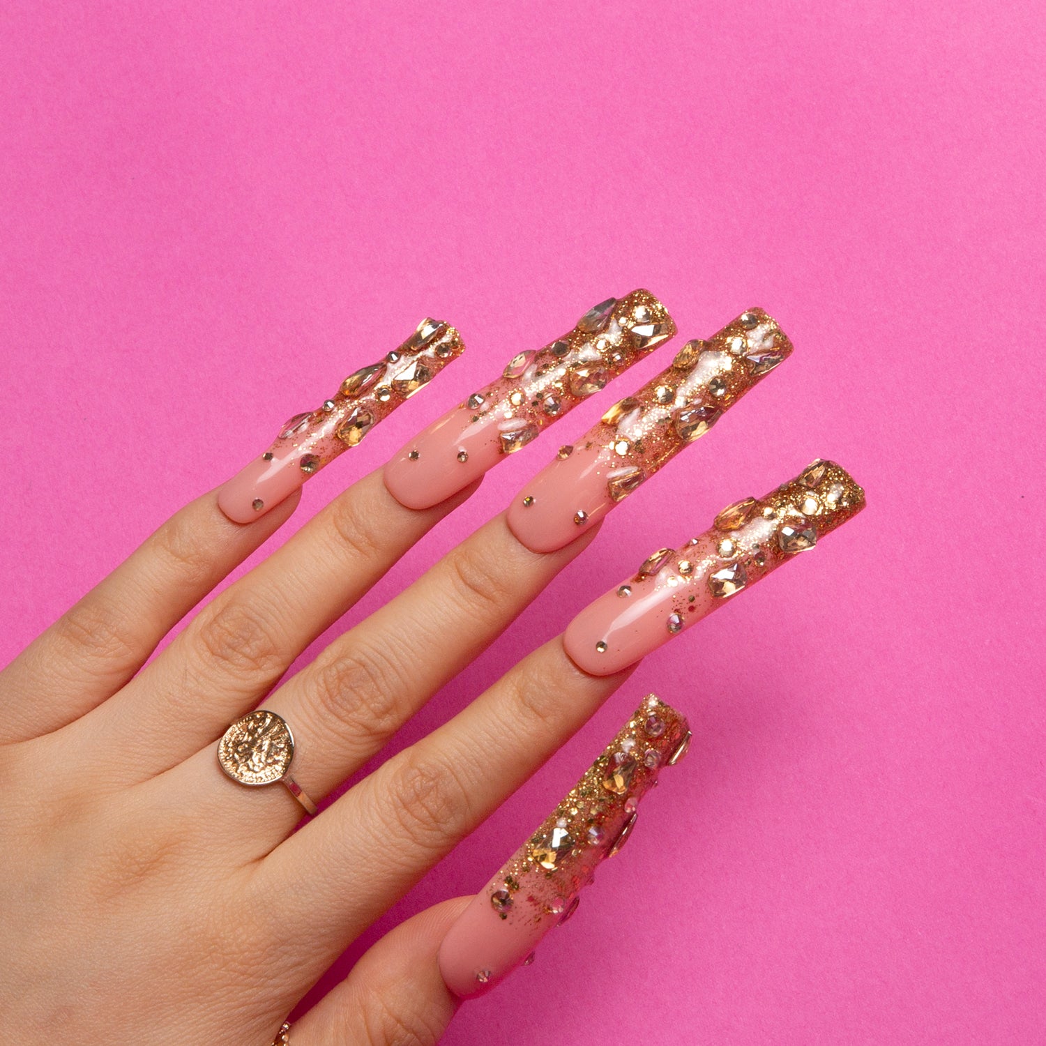 Hand with long pink and gold glittering press-on nails, ring on middle finger with a leafy pattern, on a bright pink background